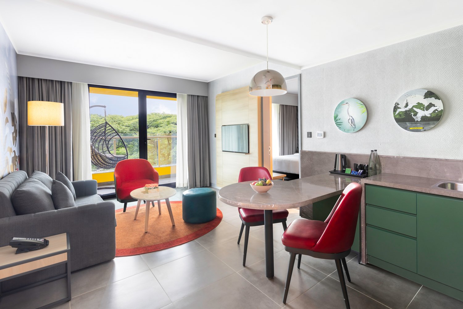 Interior photography of a modern hotel suite living area featuring a grey sofa, red armchairs, a dining table, green cabinetry, and a large window with nature views.