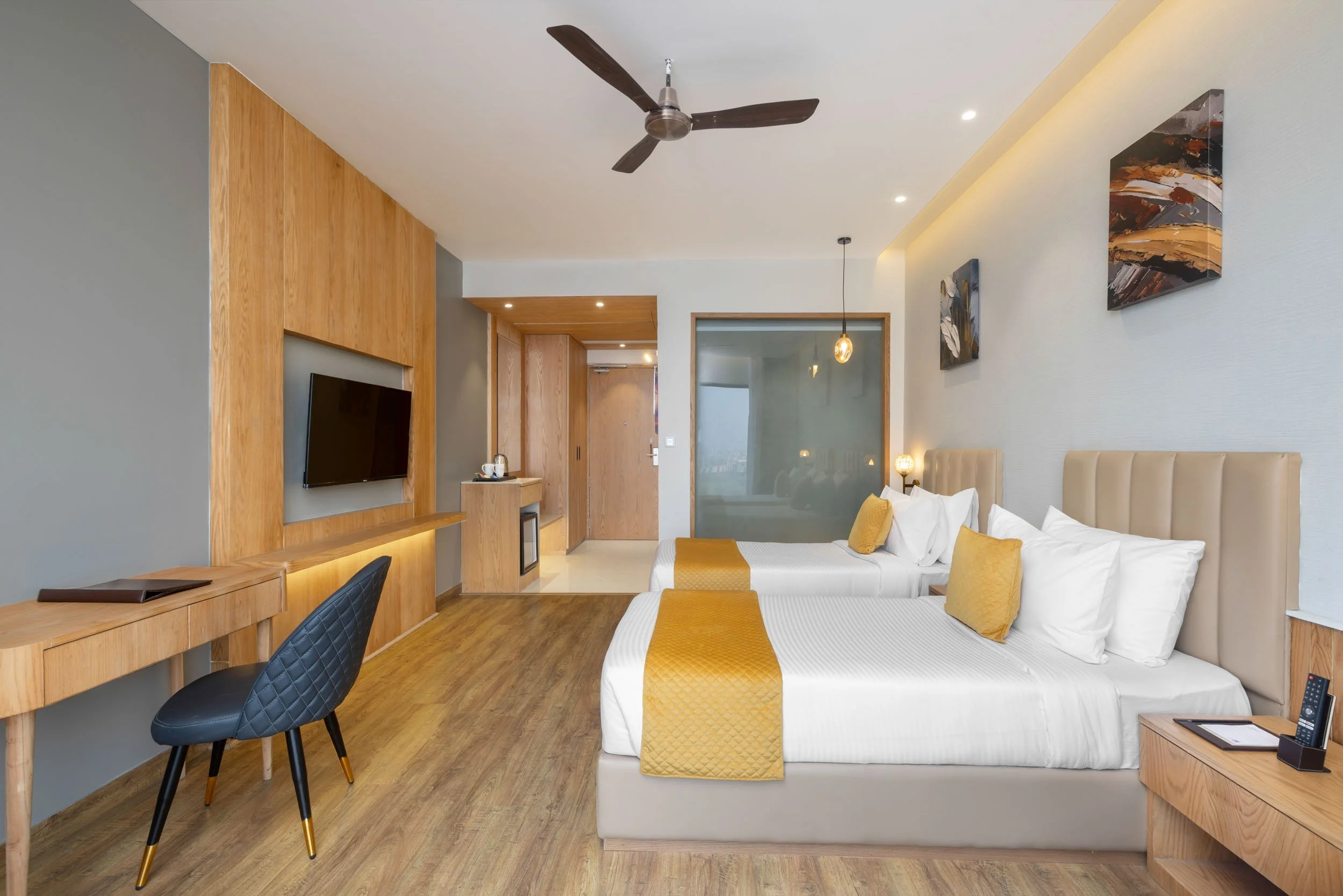 Interior photography of a modern hotel guestroom featuring a bed with a yellow runner, warm wood wall paneling, a sleek work desk with a blue chair, and natural window light
