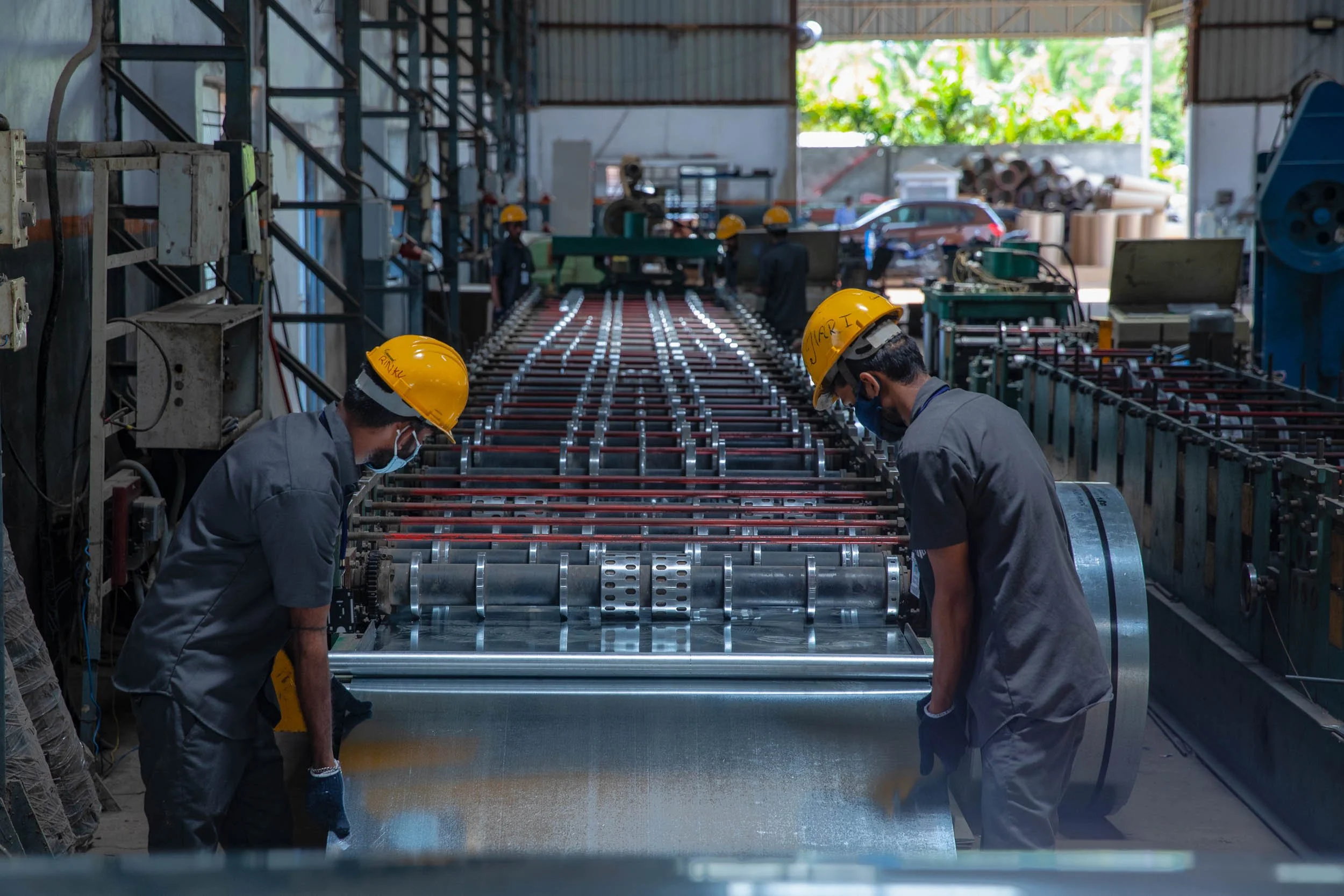 Interior industrial photography of two factory workers in yellow hard hats coordinating to handle metal pipes on a heavy-duty rack in a manufacturing plant