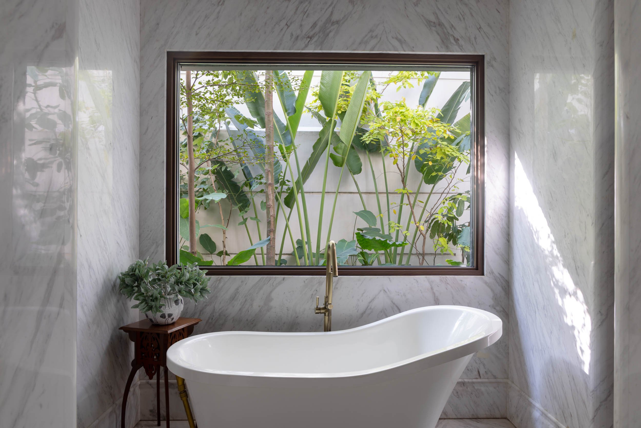 Modern bathroom design featuring a freestanding white bathtub positioned next to a large window framing lush tropical plants