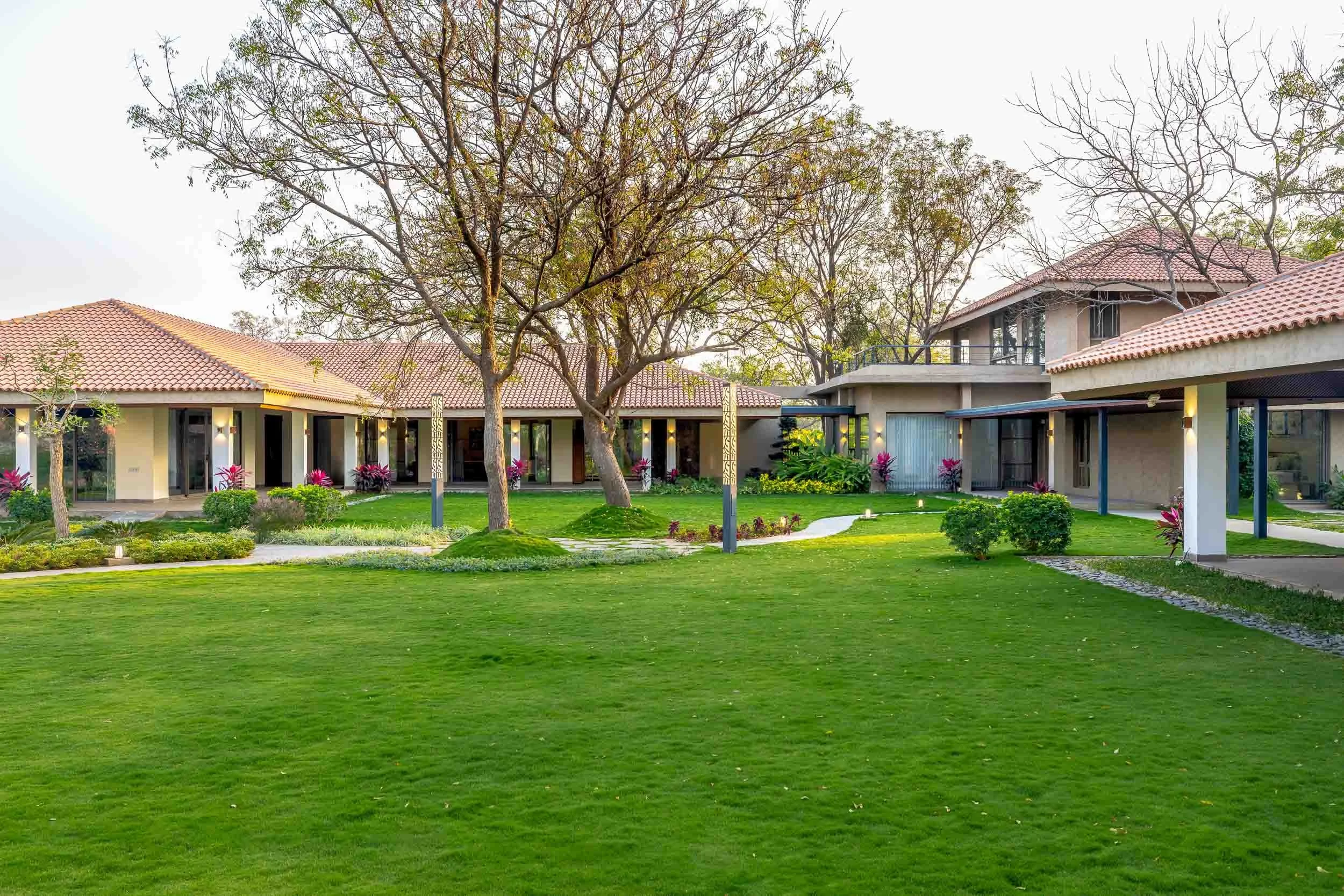 Exterior daytime photography of a luxury resort farm stay featuring an expansive green lawn, a large mature tree, and a long traditional building with a pitched roof and veranda
