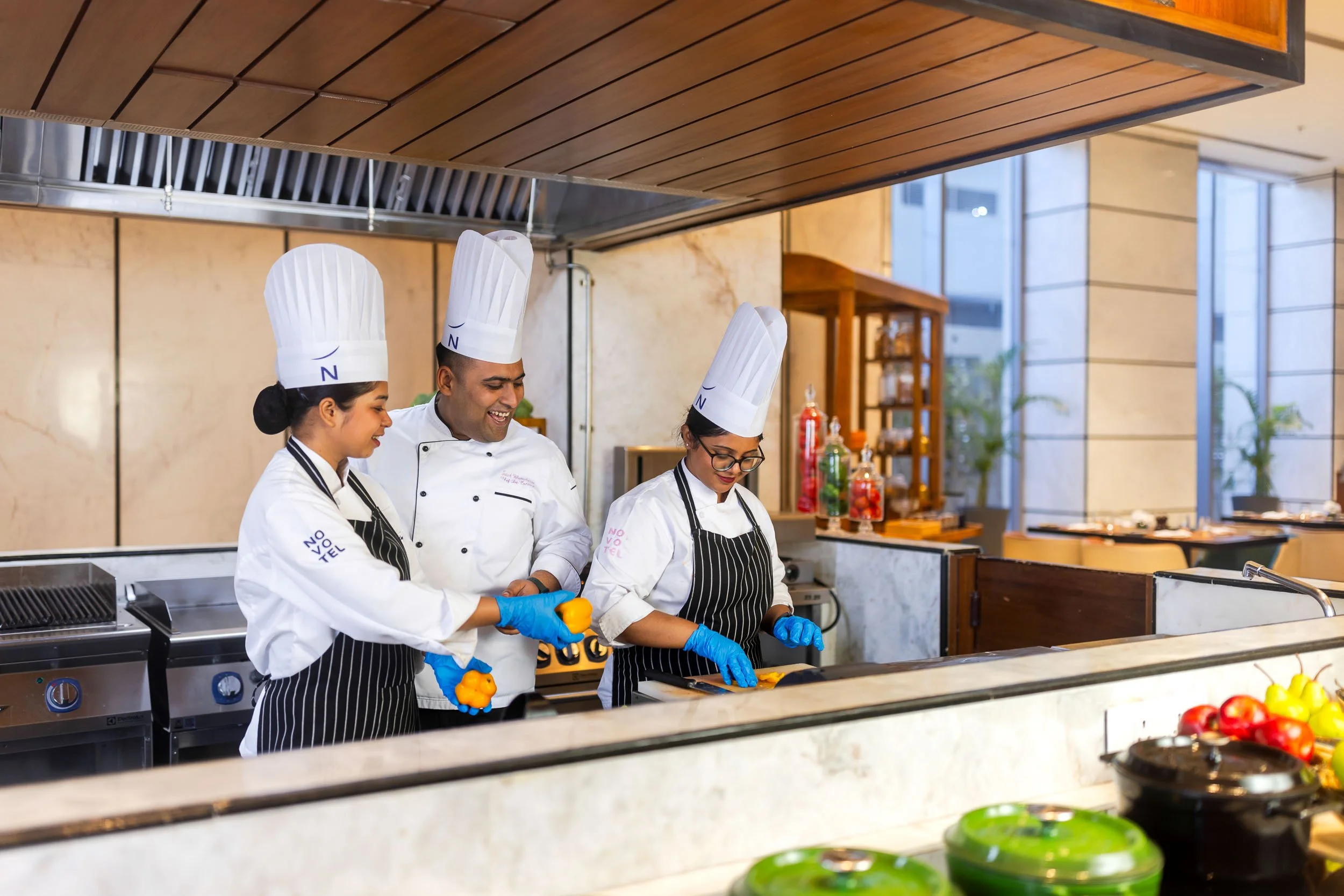 Interior photography of a modern hotel live cooking station featuring three chefs in uniform preparing food, a wooden slatted overhead design, and fresh vegetables on display