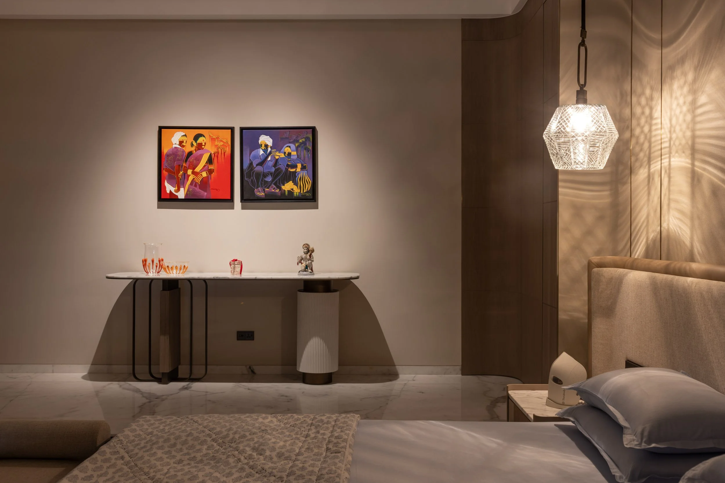 Modern residential bedroom interior featuring warm ambient lighting, a bed, a wooden console, two colorful framed artworks, and a pendant light