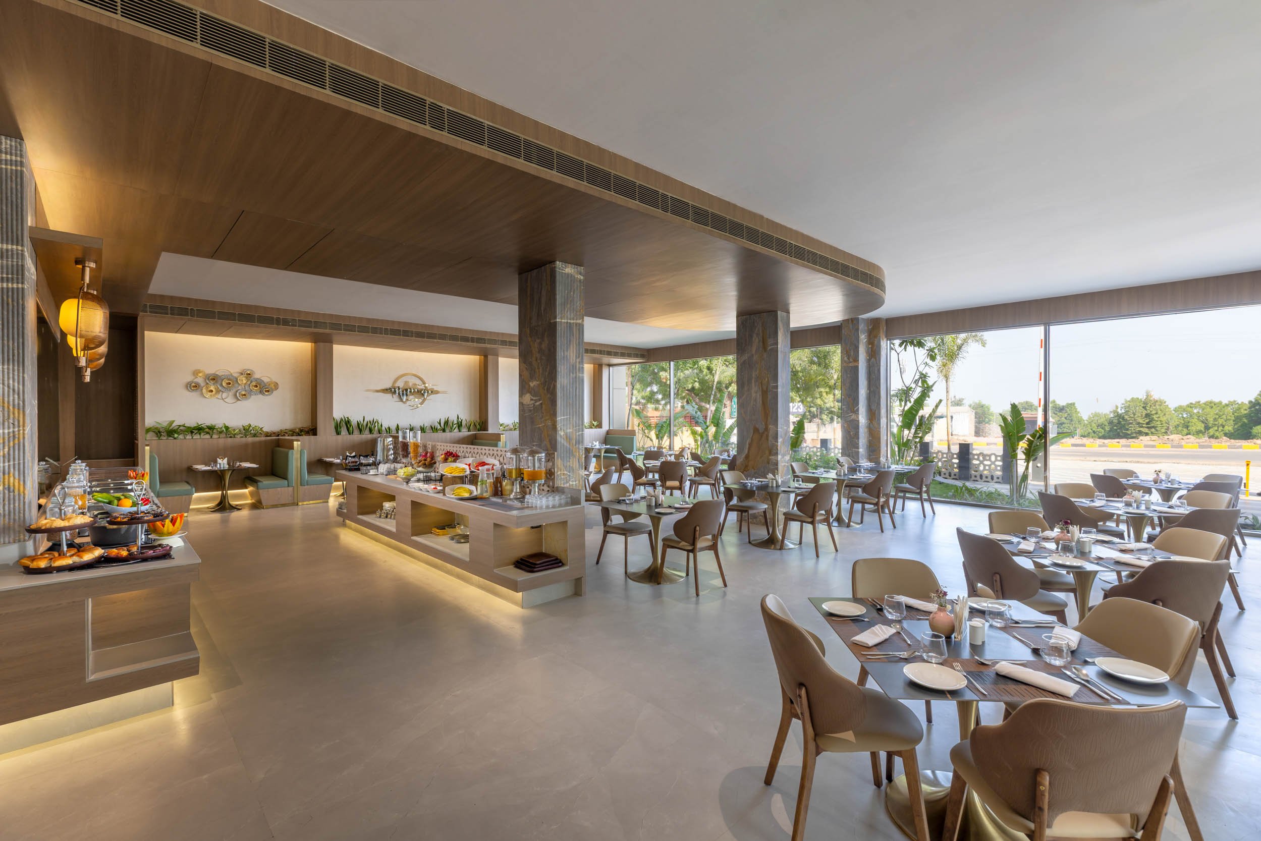 Interior photography of a spacious modern hotel restaurant featuring wooden dining tables and chairs, buffet stations, and bright ambient lighting