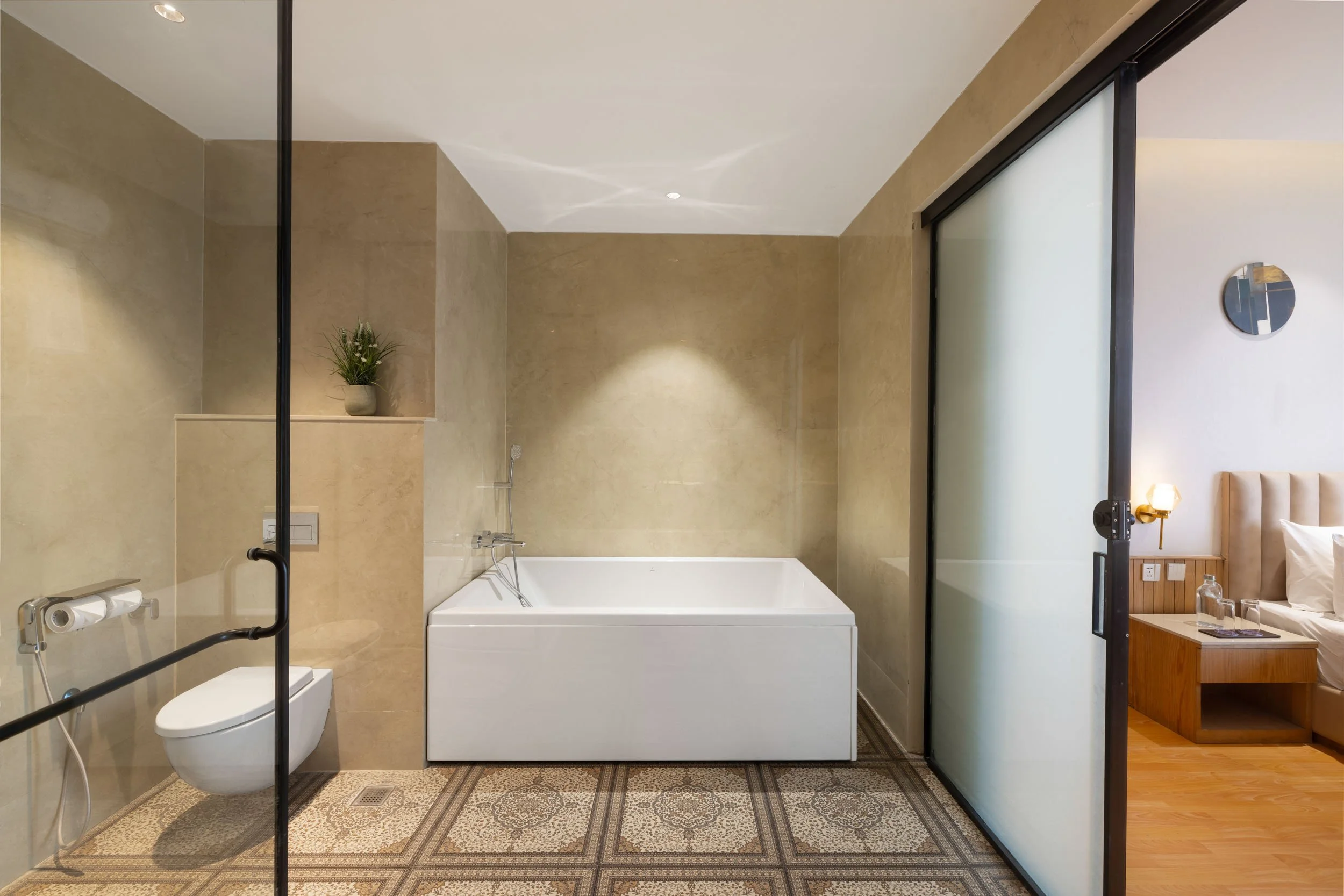 Interior photography of a modern luxury hotel bathroom featuring a white soaking bathtub, beige textured wall tiles, a glass shower enclosure, and warm ambient lighting.