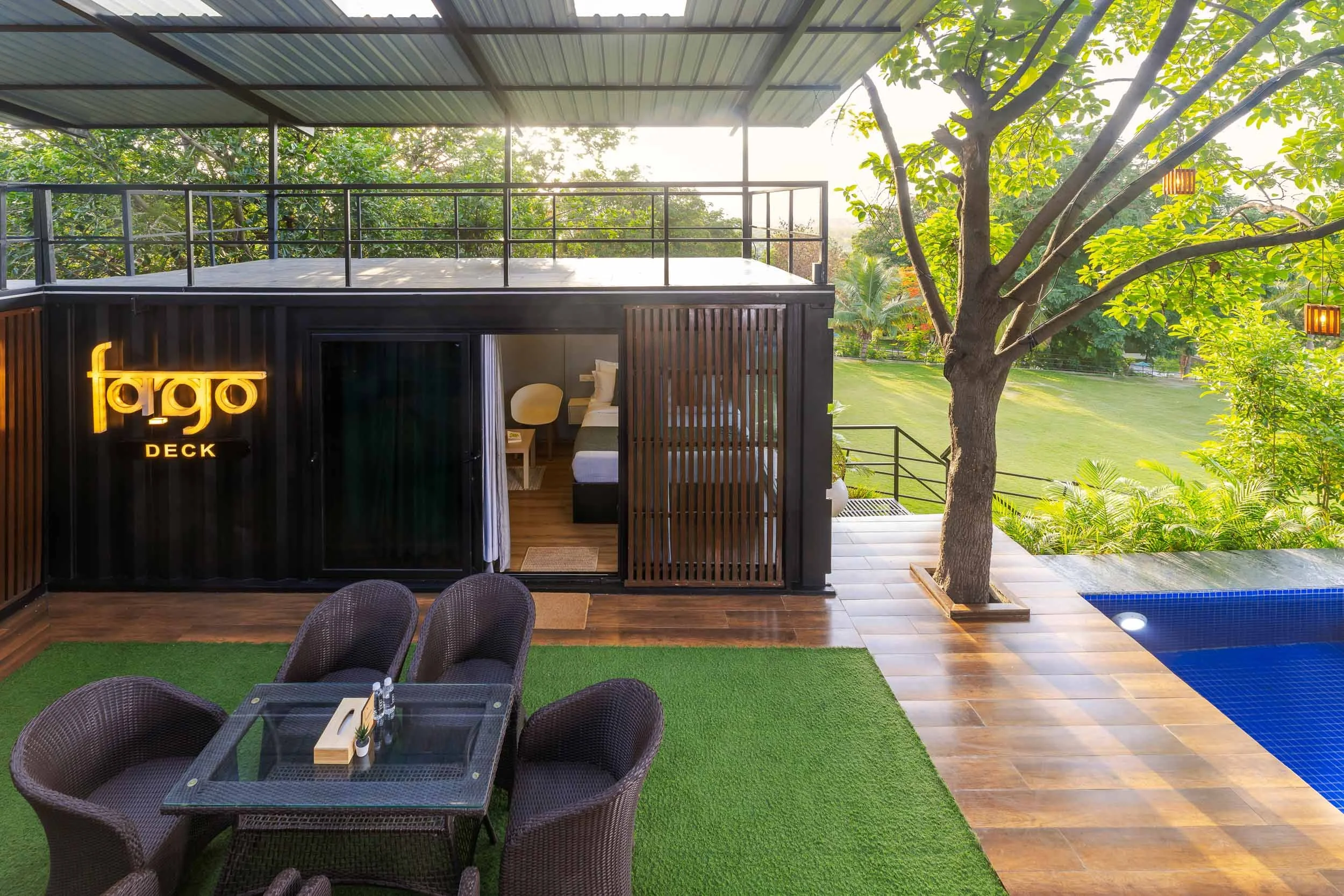 Exterior daytime photography of a modern luxury staycation in Hyderabad, featuring a covered outdoor deck with wicker seating, wooden slatted privacy screens, and garden views