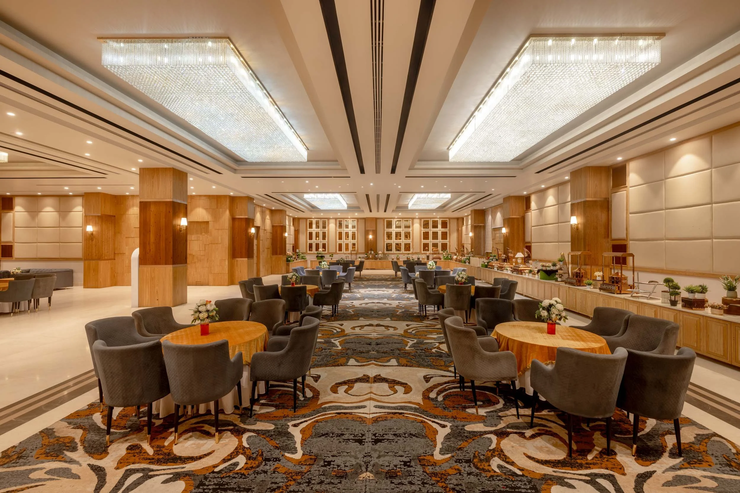 Interior photography of a luxury hotel restaurant featuring round dining tables, dark upholstered chairs, a patterned carpet, and a striking recessed wooden ceiling with large light panels