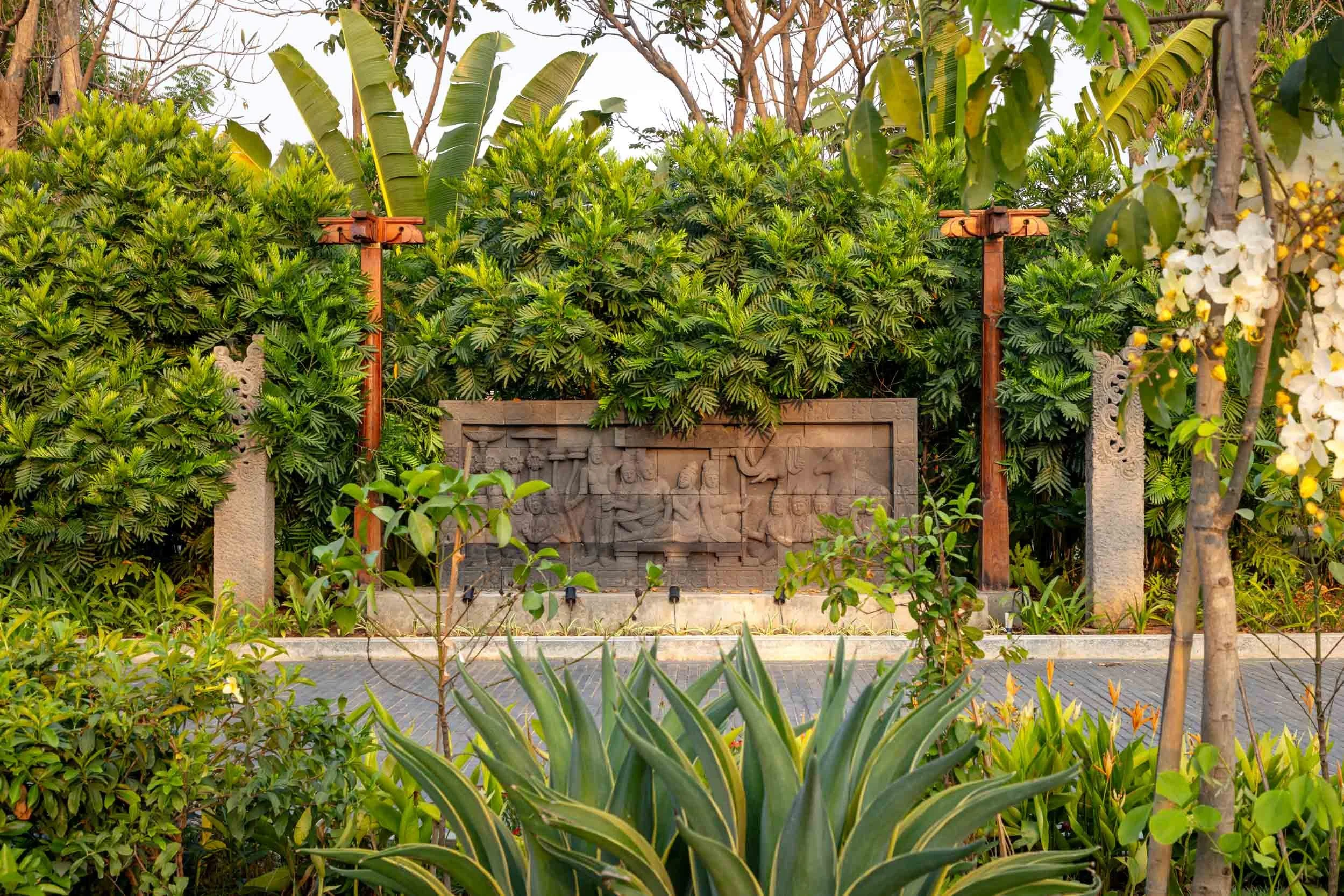 Residential landscape photography featuring a textured stone feature wall flanked by wooden lampposts and lush tropical greenery, including large agave plants.
