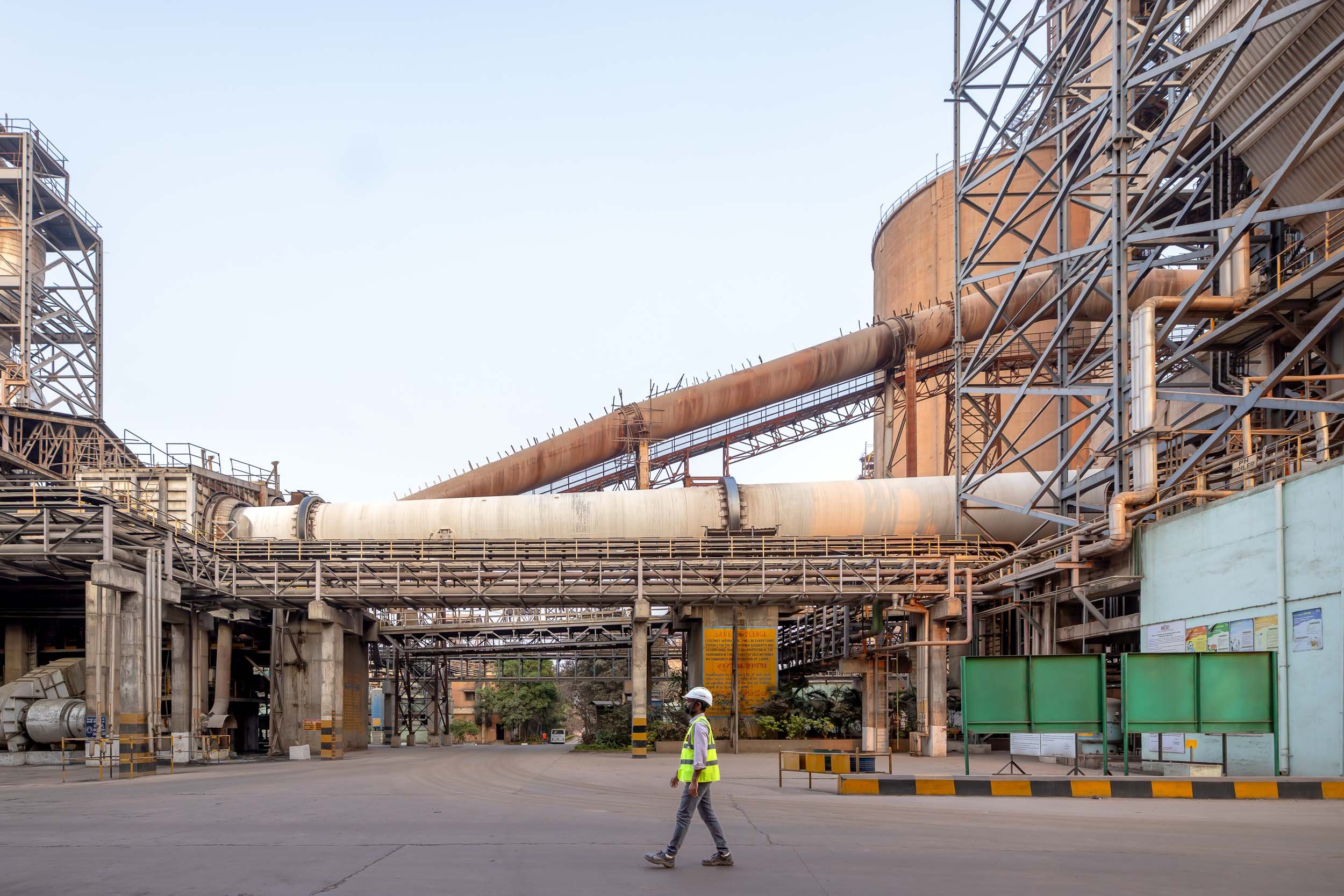 Wide exterior photography of a large-scale industrial manufacturing plant in India, featuring heavy machinery, complex steel structures, and a worker in safety gear