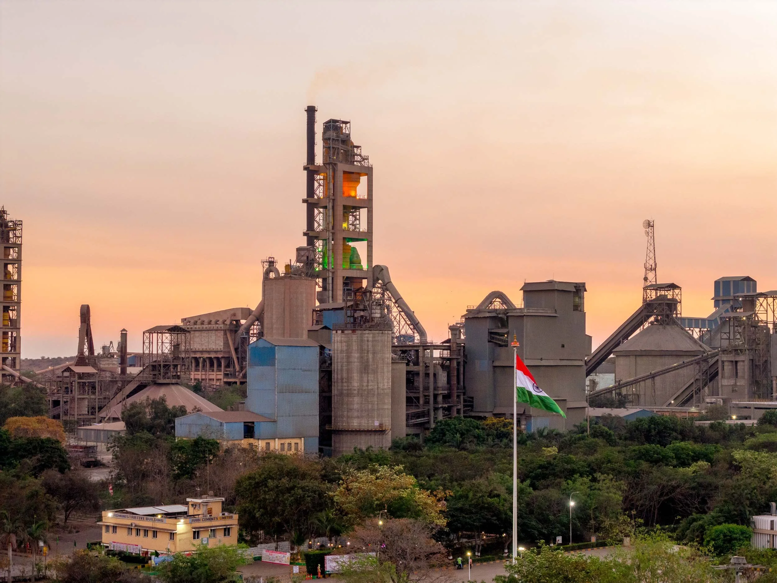 Exterior twilight photography of a large-scale industrial manufacturing plant in India, featuring tall silos, factory infrastructure, and the Indian flag at sunset