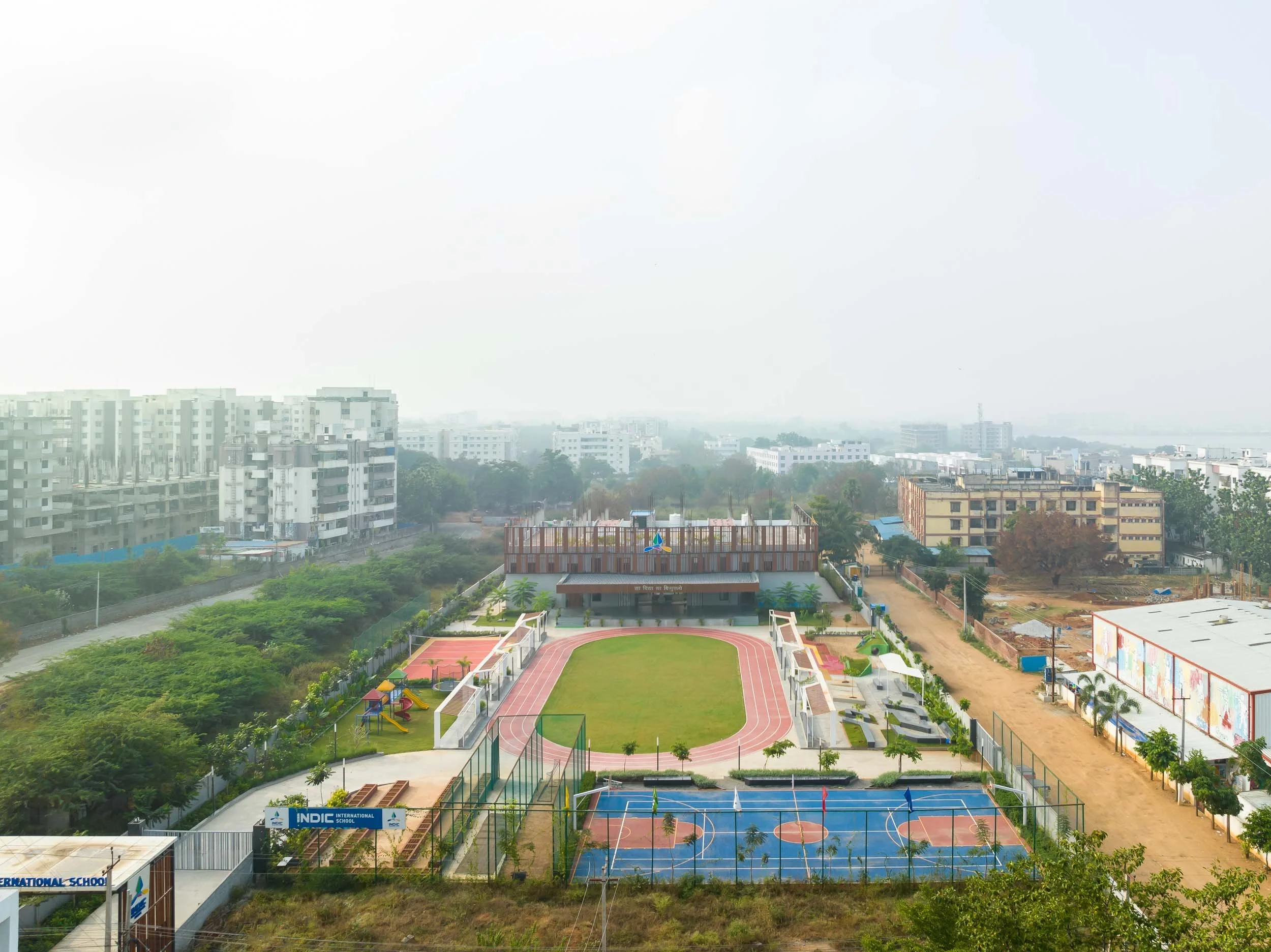 An aerial bird's-eye view of a large school campus, featuring multi-story buildings, lush green surroundings, and a central courtyard with bright blue and red outdoor basketball and tennis courts