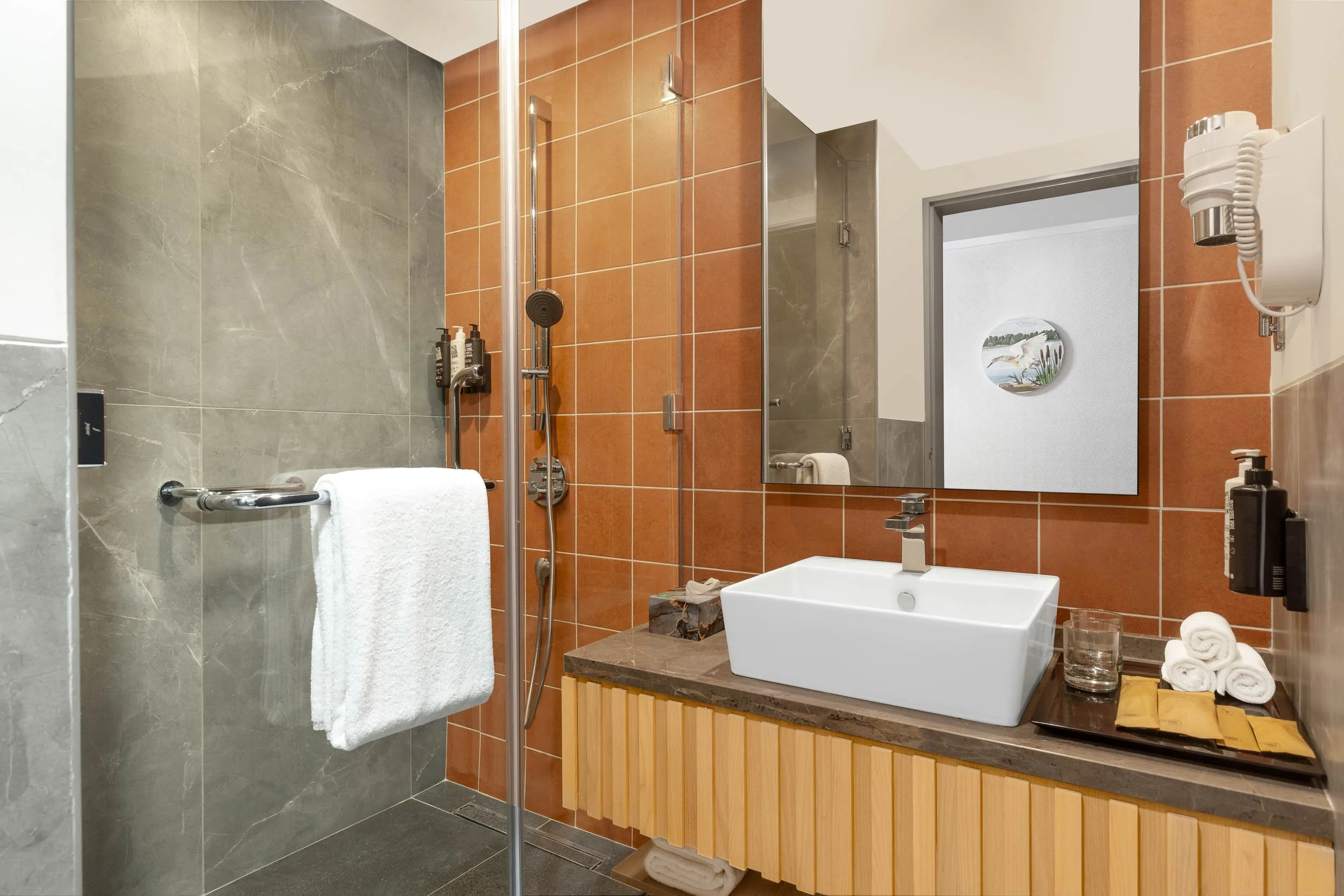 Interior photography of a modern hotel bathroom featuring a white vessel sink on a wooden vanity, a large mirror, and sleek grey tiled walls