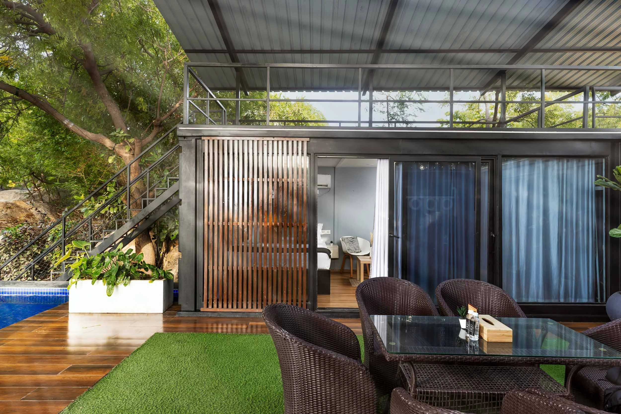 Exterior daytime photography of a modern luxury staycation cabin in Hyderabad, featuring a covered outdoor deck with wicker seating, a green turf lawn, and wooden slatted privacy screens
