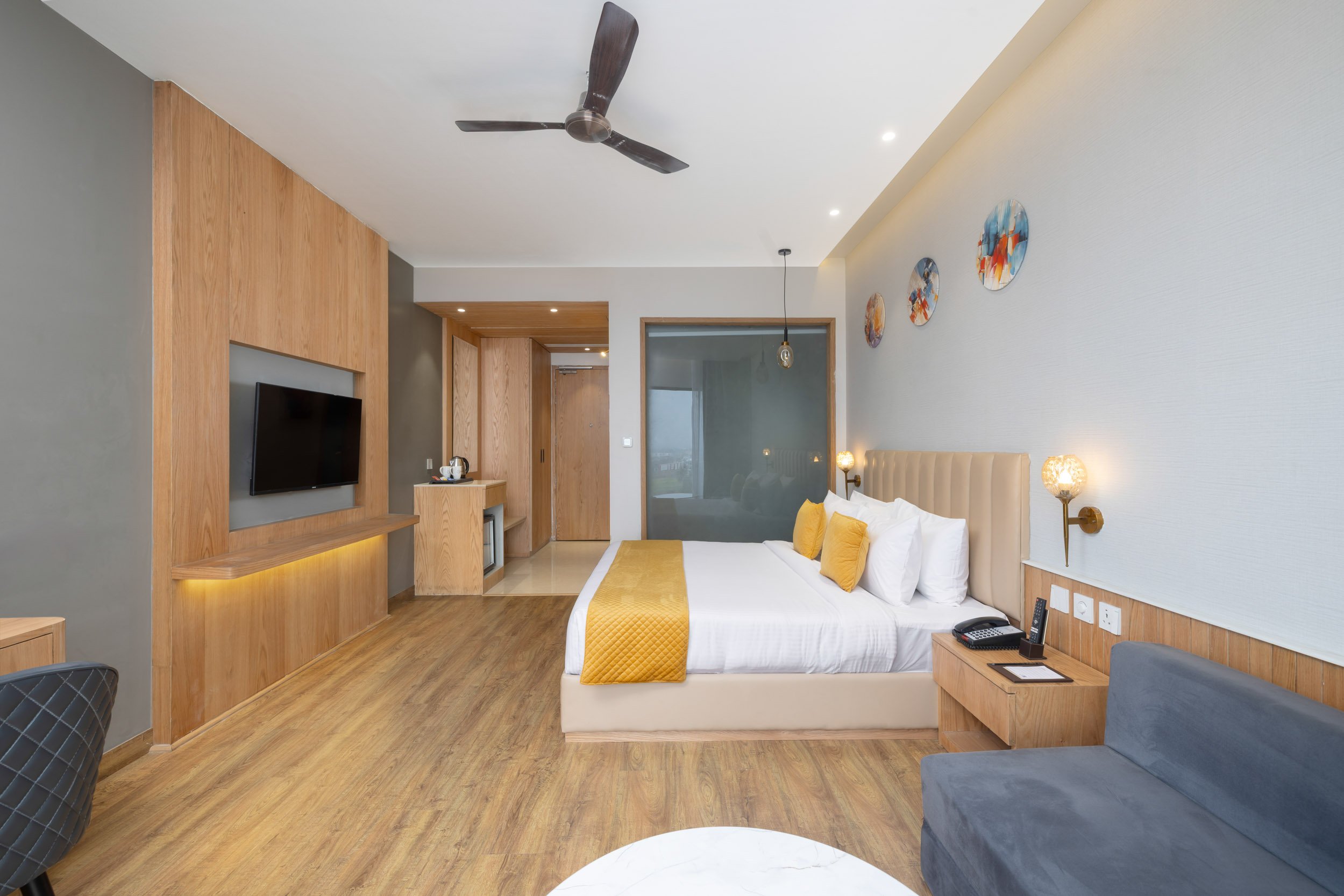 Interior photography of a spacious modern hotel room featuring a king bed with a yellow runner, wood wall paneling, a comfortable blue sofa, and a wall-mounted TV