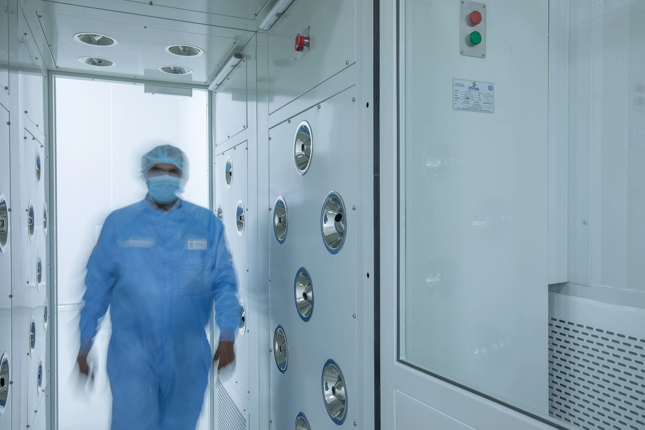 Interior industrial photography of a worker in blue sterile gear walking through a clean room corridor lined with control panels in a manufacturing facility.
