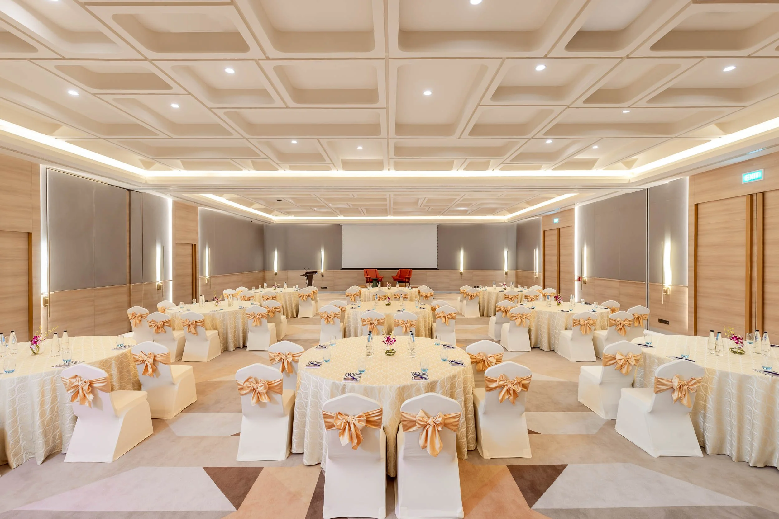 Interior photography of a spacious hotel banquet hall featuring round tables with white chair covers and orange bows, a patterned carpet, an elaborate ceiling with warm lighting, and a large projection screen