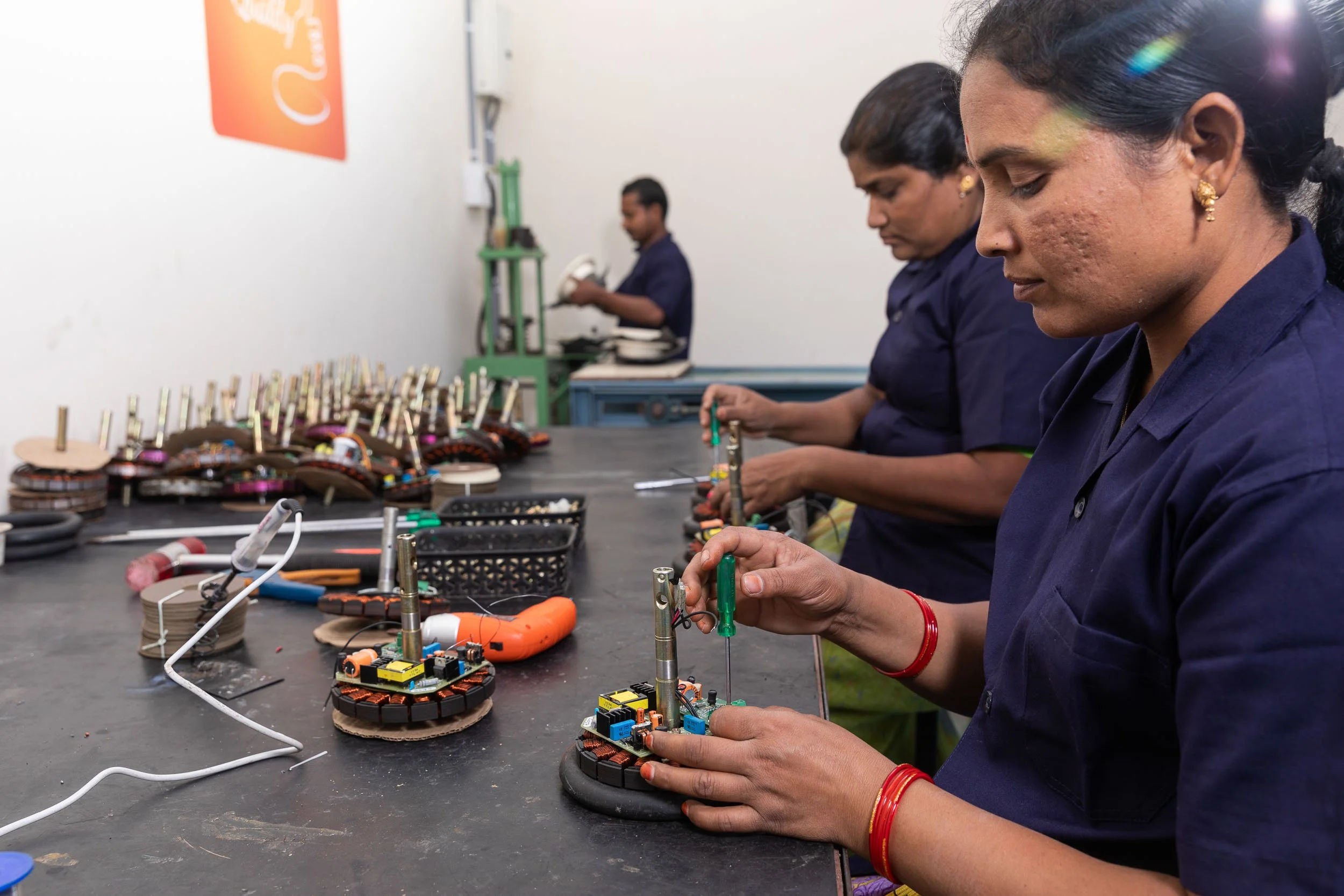 Interior industrial photography of female factory workers in blue uniforms carefully assembling intricate components on a production line in a manufacturing plant