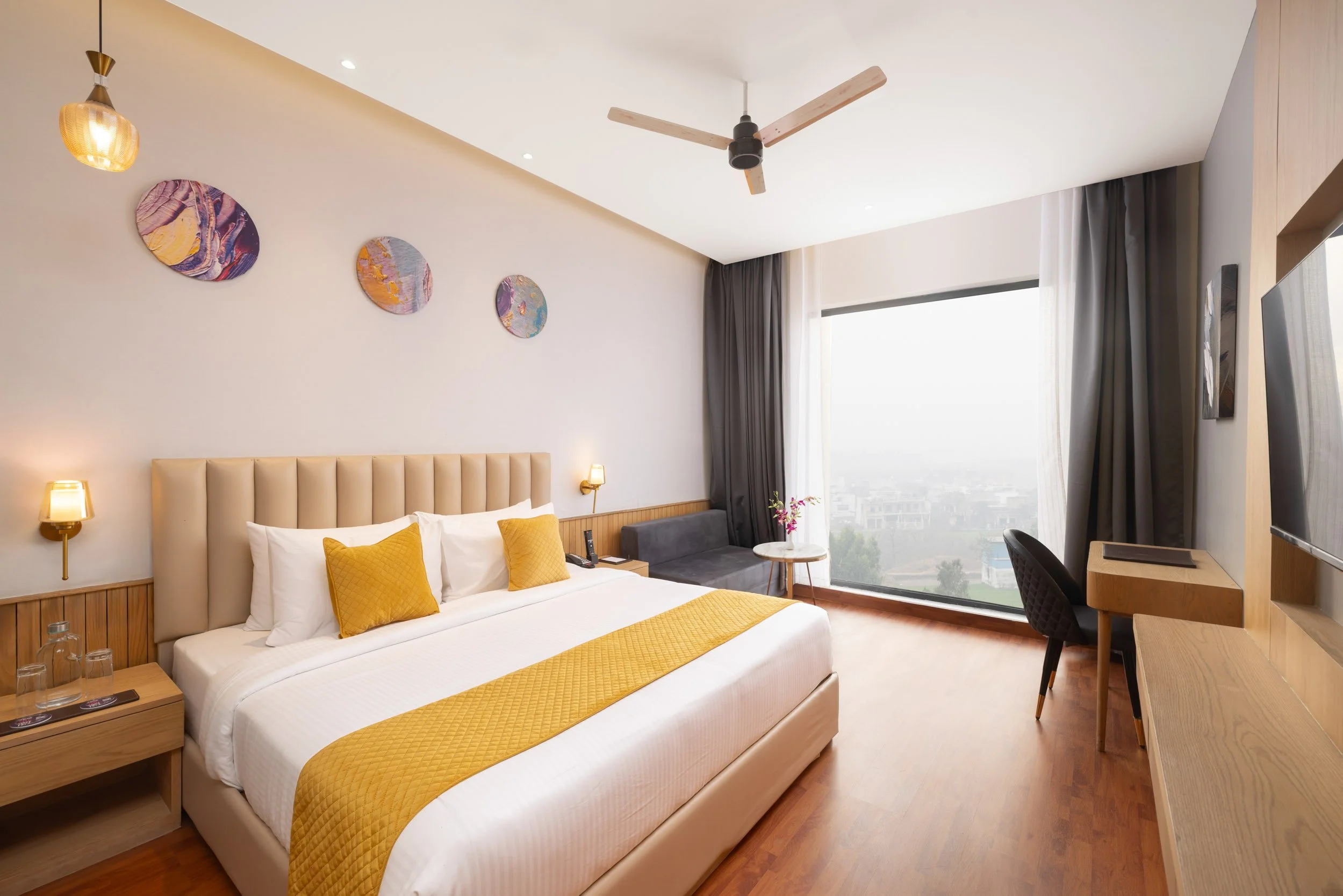 Interior photography of a modern hotel king room featuring a bed with a yellow runner, circular wall art, wooden flooring, and a window with a dedicated workspace