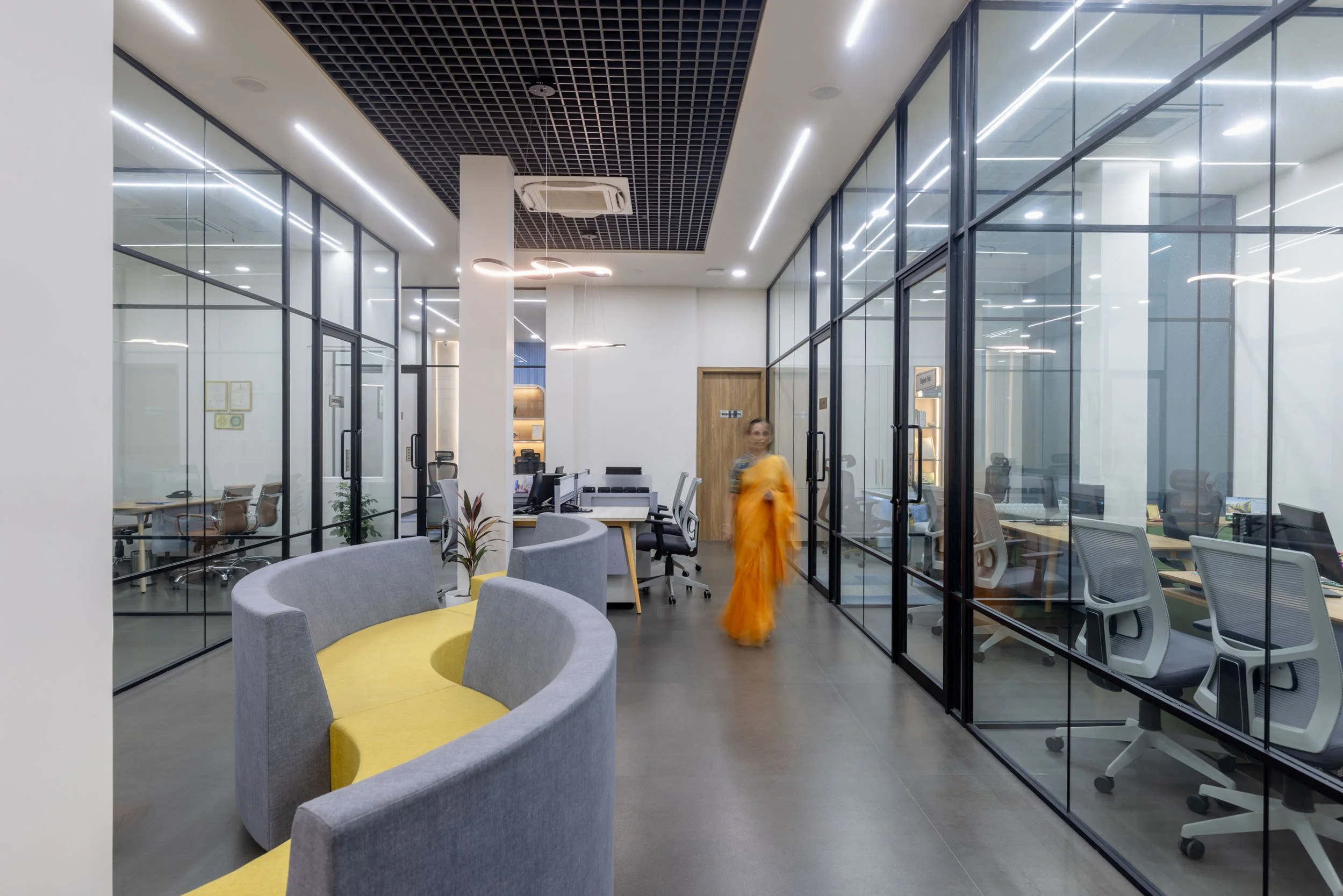 Commercial interior photography showcasing a dynamic office hallway, highlighting transparent glass-enclosed workspaces, dark industrial ceilings, and vibrant yellow lounge seating