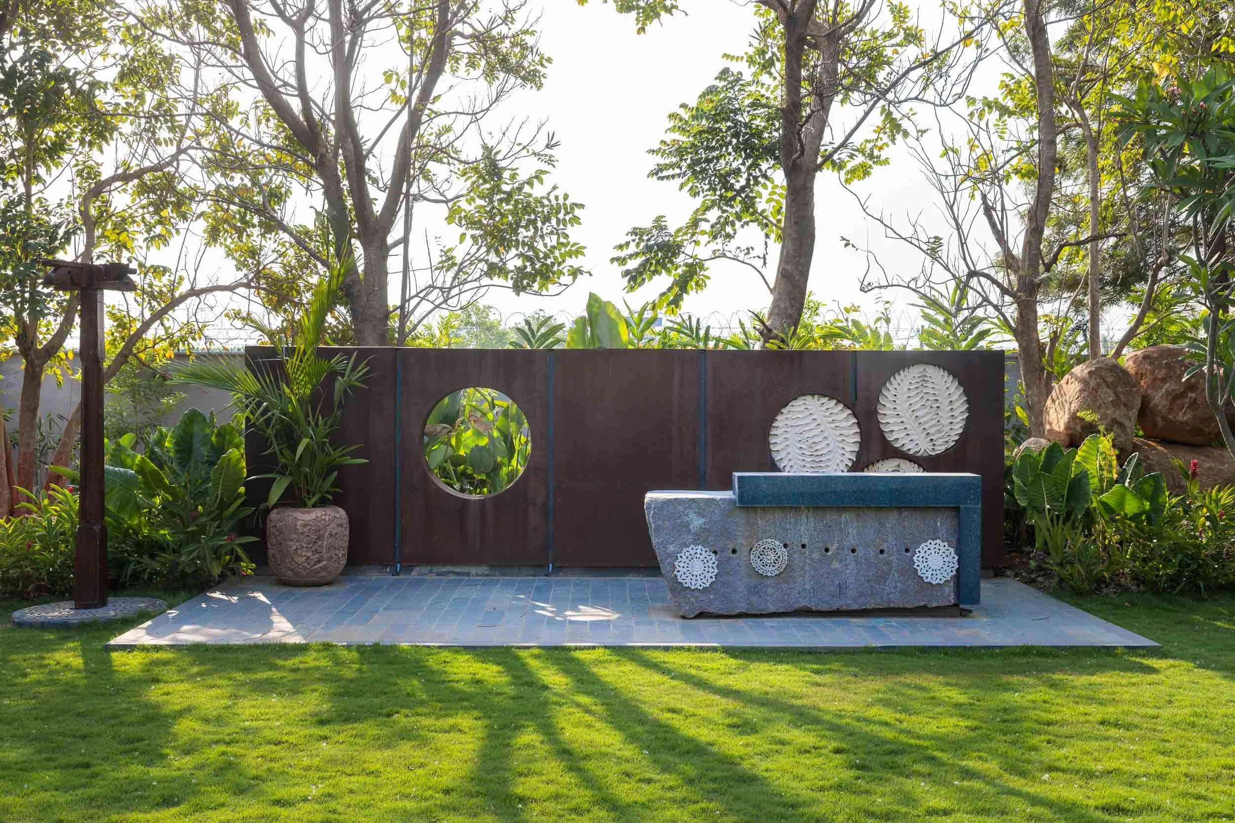 Exterior daytime photography of a modern landscape feature at a luxury resort, showing a green lawn, a dark textured wall with a circular cutout, minimalist outdoor seating, and tall trees