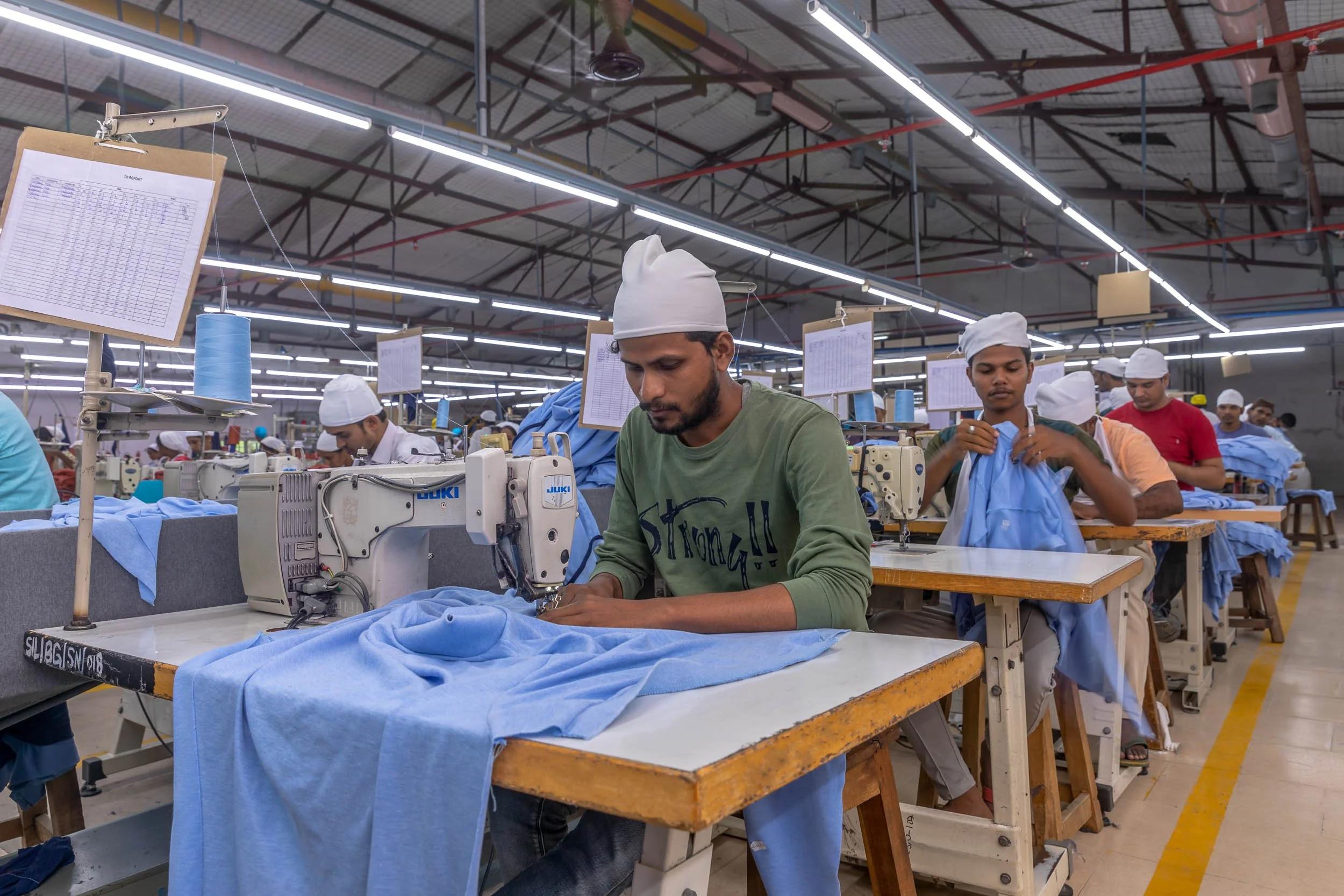 Interior industrial photography of garment workers sewing light blue fabric at workstations in a large-scale textile manufacturing plant in India