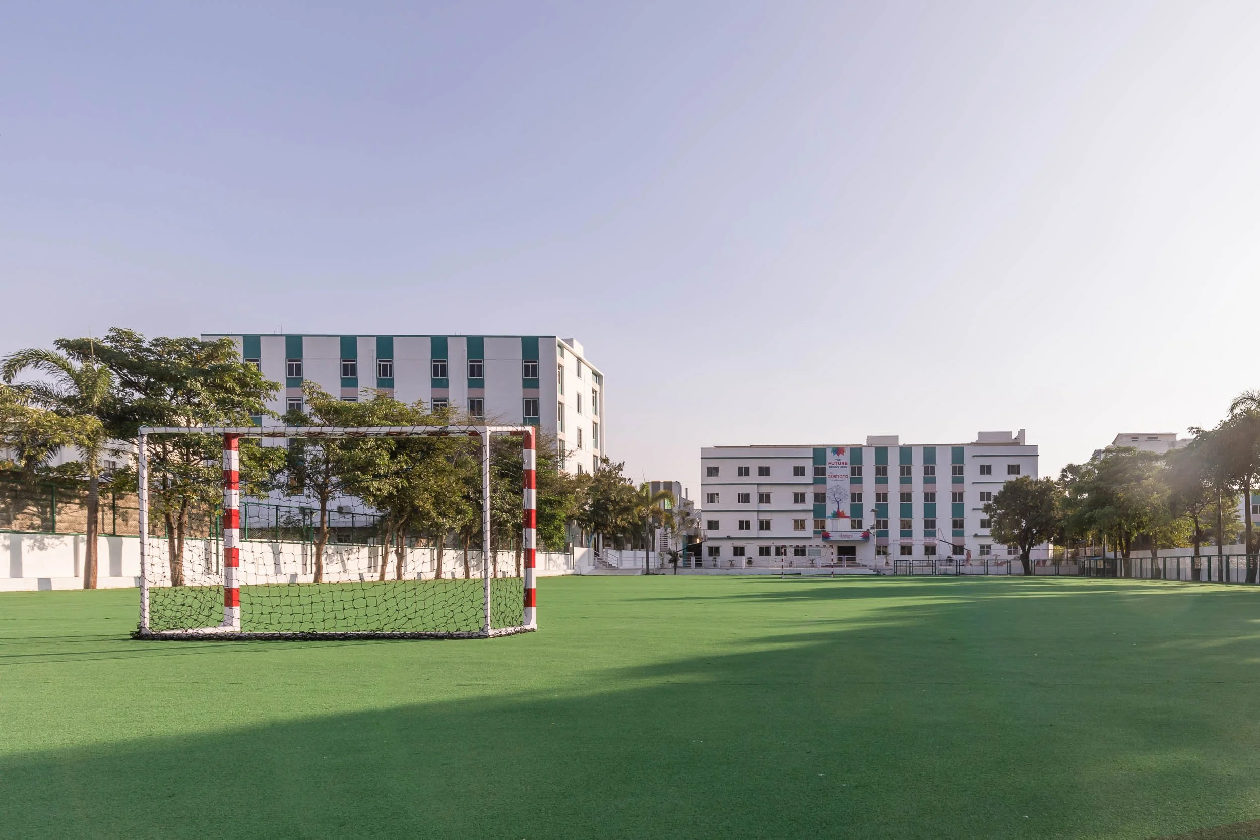 A wide exterior photo of a green artificial turf sports field with a white soccer goal post on the left, backed by a large, modern, multi-story white school building