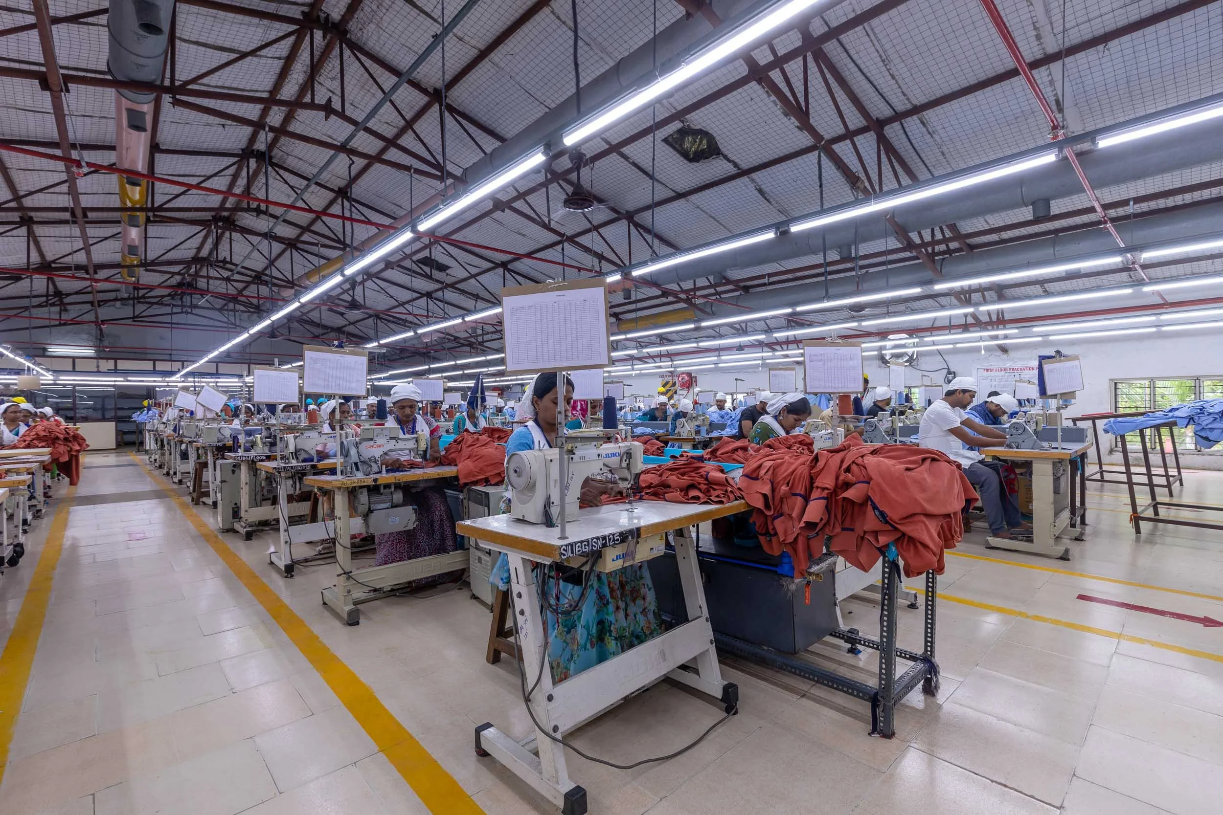 Wide interior industrial photography of a brightly lit textile manufacturing plant floor, featuring long assembly lines, workers at their stations, and a yellow safety line
