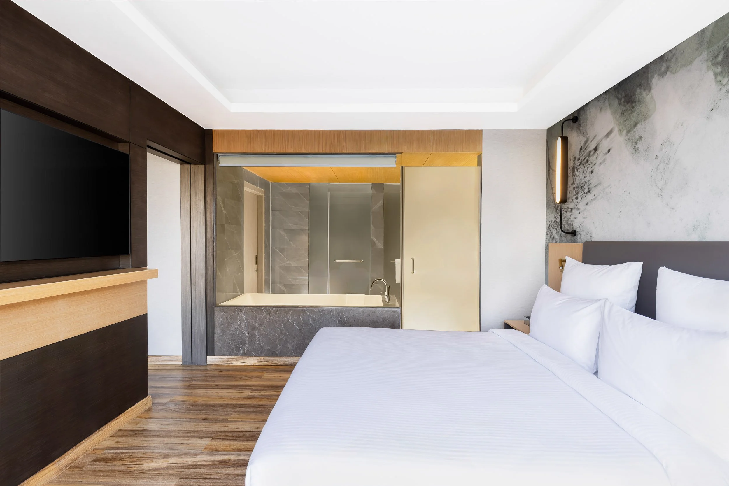 Interior photography of a modern hotel bedroom featuring a large white bed, wooden flooring, a wall-mounted TV on a dark accent panel, and contemporary artwork
