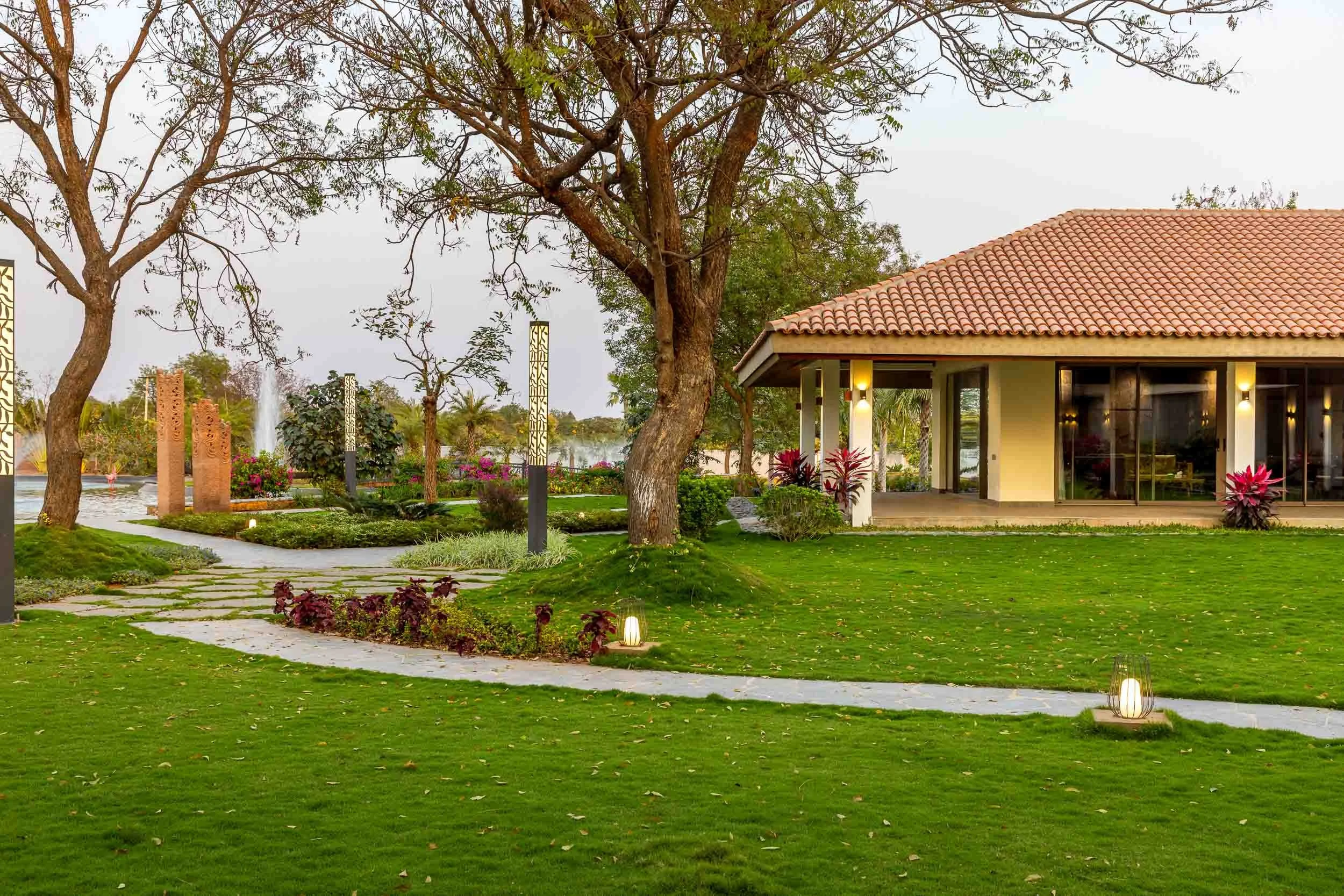 Exterior daytime photography of a luxury resort farm stay featuring a sweeping green lawn, a winding stone pathway, mature trees, and a traditional pitched-roof building