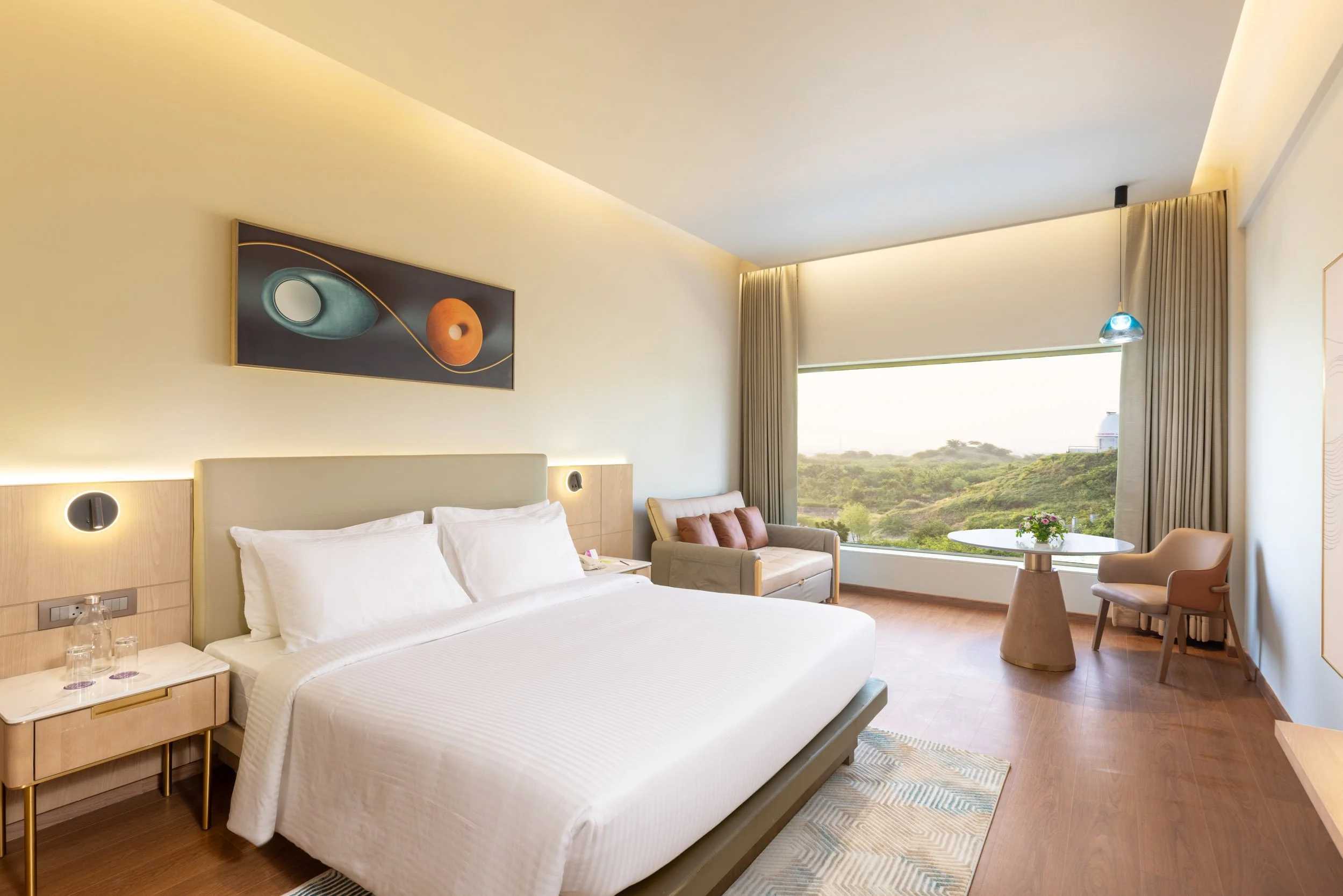 Interior photography of a modern hotel king room featuring a bed with a wooden headboard, contemporary horizontal wall art, and a seating area by a large window