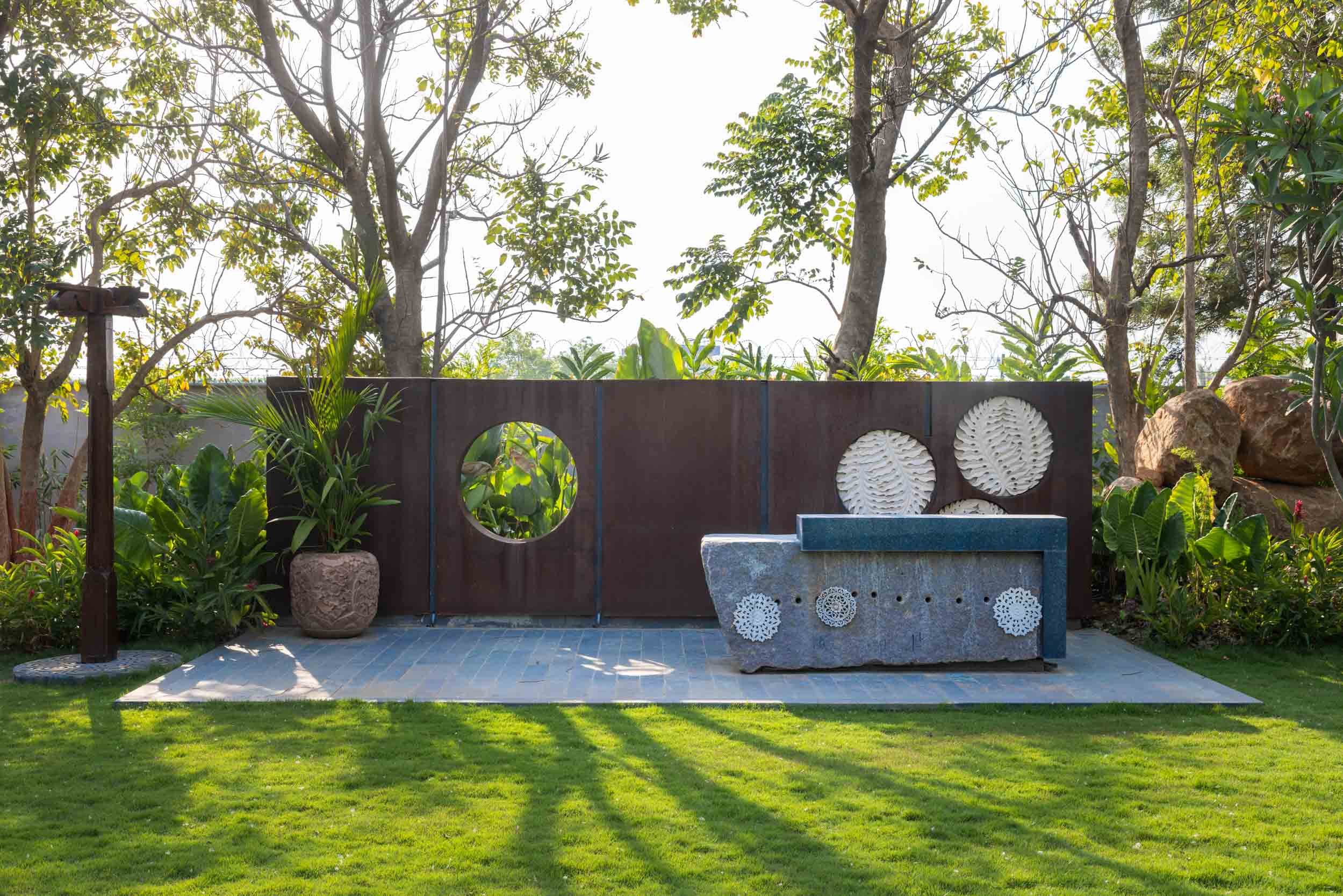 Modern residential landscape featuring a sunlit, manicured lawn and a custom dark wood slatted boundary wall with circular cutouts and built-in seating.