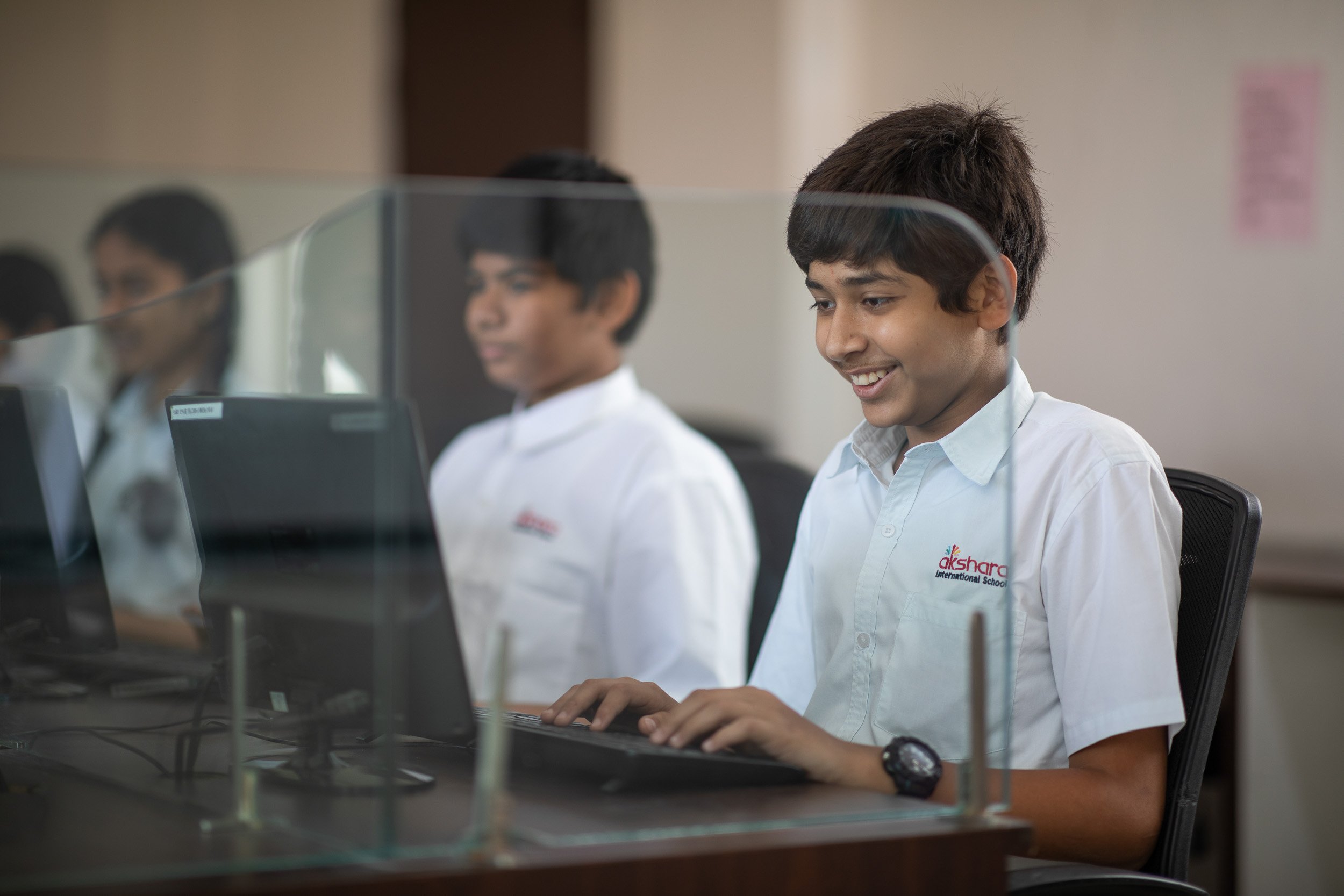Three students in white school uniforms sitting at desks in a brightly lit computer lab, smiling as they look together at their desktop monitors