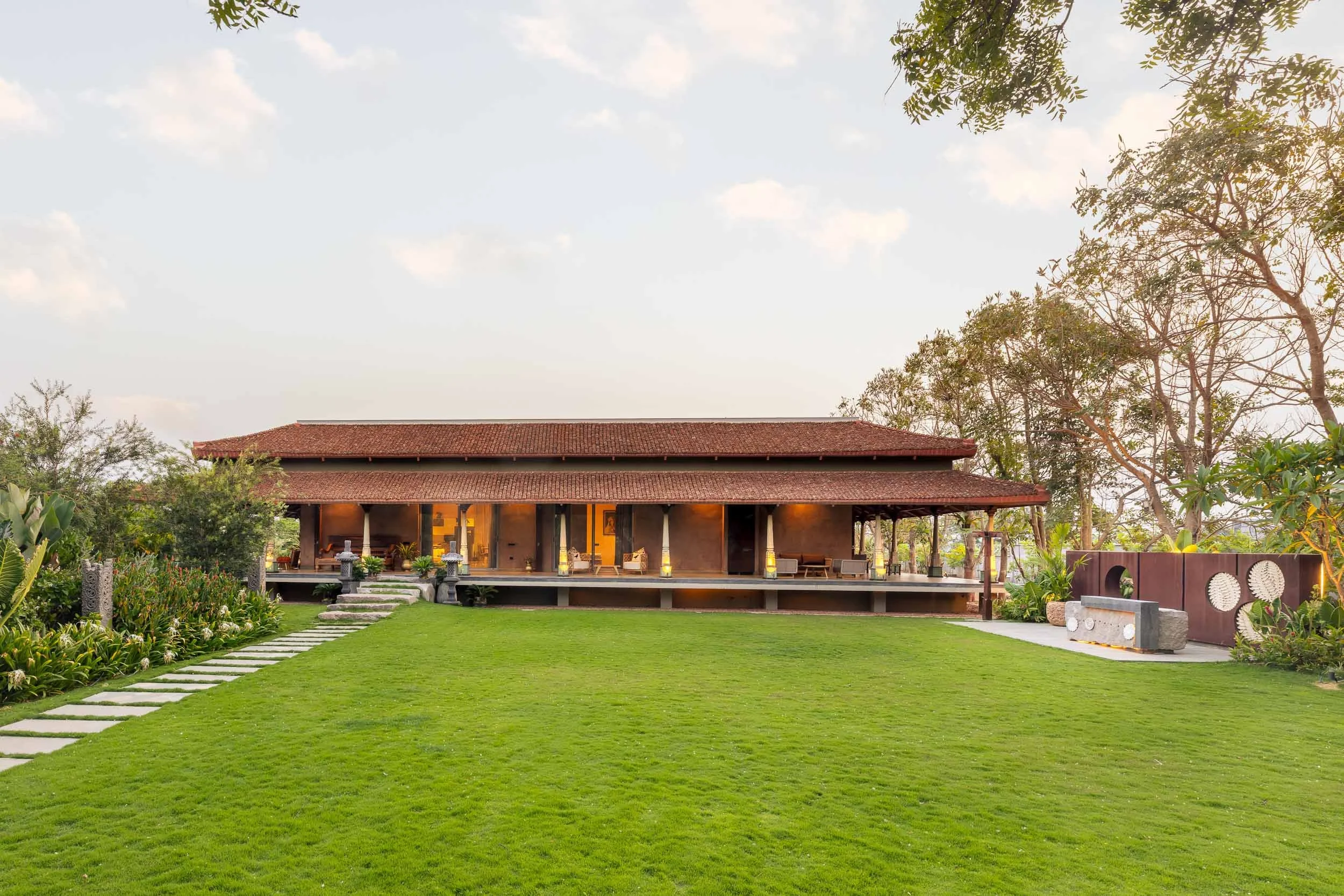 Exterior daytime photography of a luxury resort farm stay featuring a lush garden with vibrant yellow flowering bushes and a traditional pitched-roof building in the background