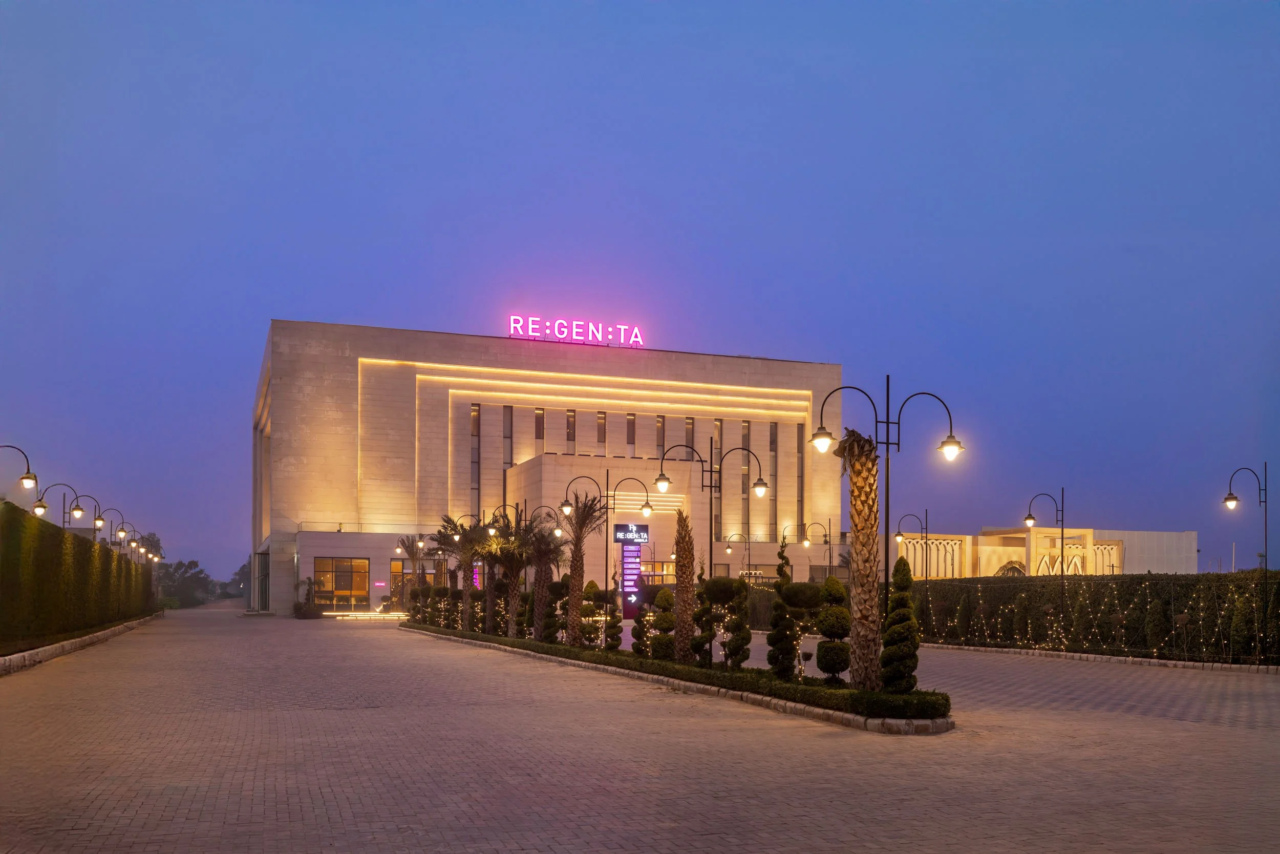 Exterior twilight photography of the Regenta hotel featuring a warmly illuminated facade, a paved driveway entrance, and decorative pathway lighting