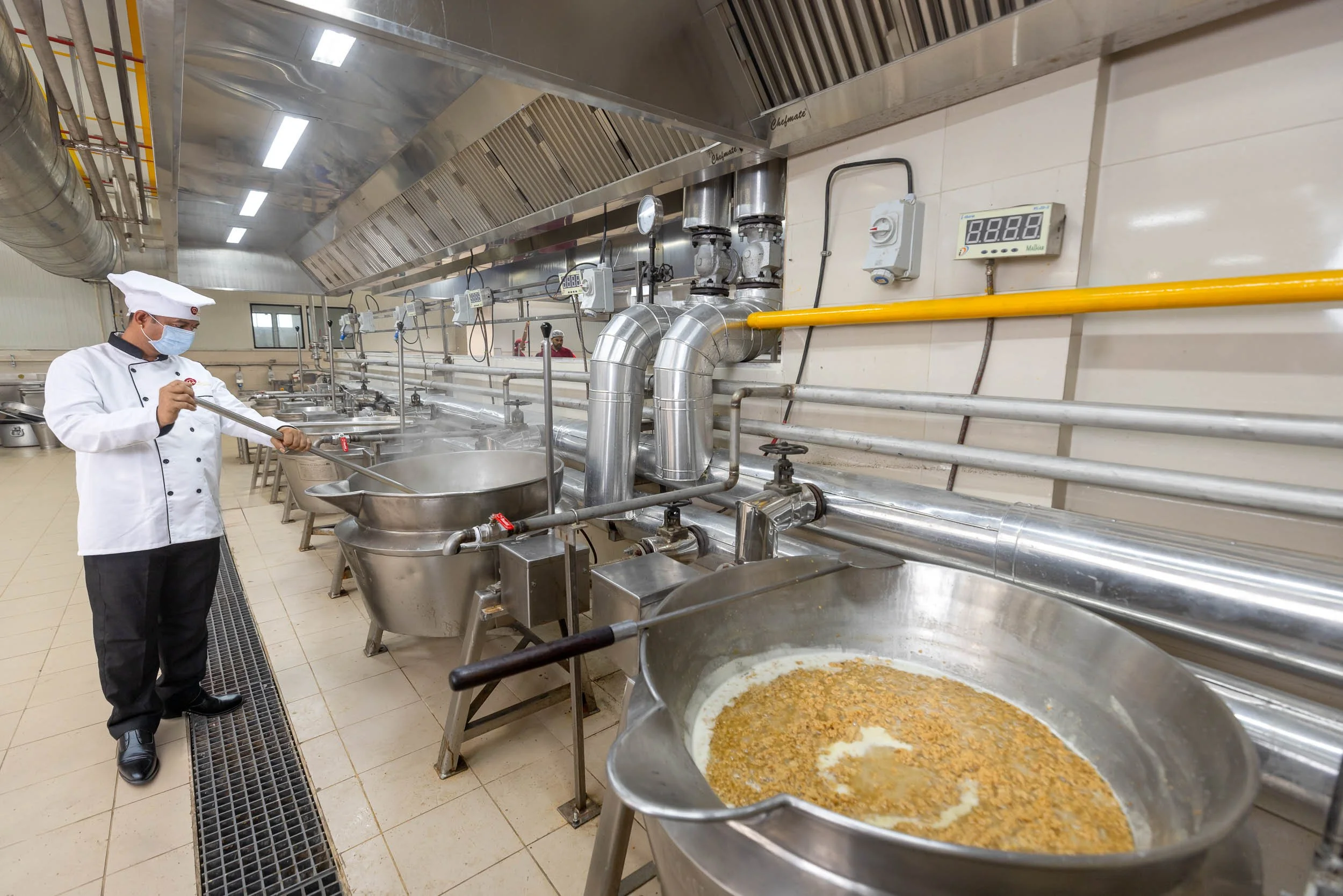 Interior industrial photography of a worker in a white uniform operating large stainless steel cooking kettles in a commercial food processing facility.