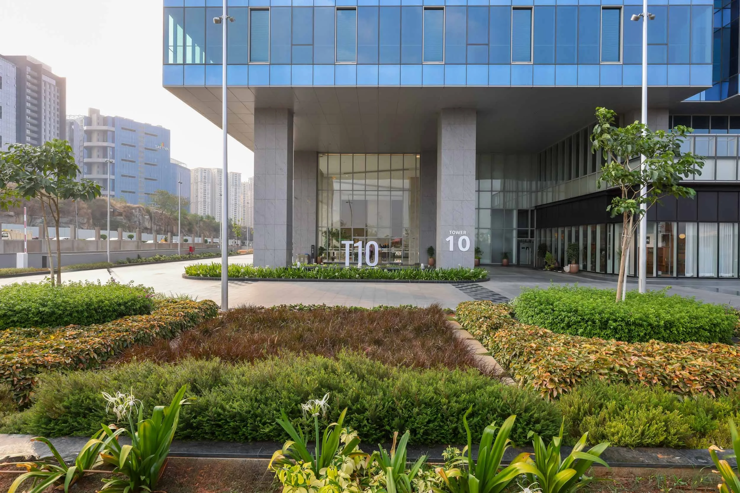 Professional commercial architecture photography capturing a ground-level perspective of a modern office building, highlighting a beautifully landscaped entrance and a sleek glass facade.