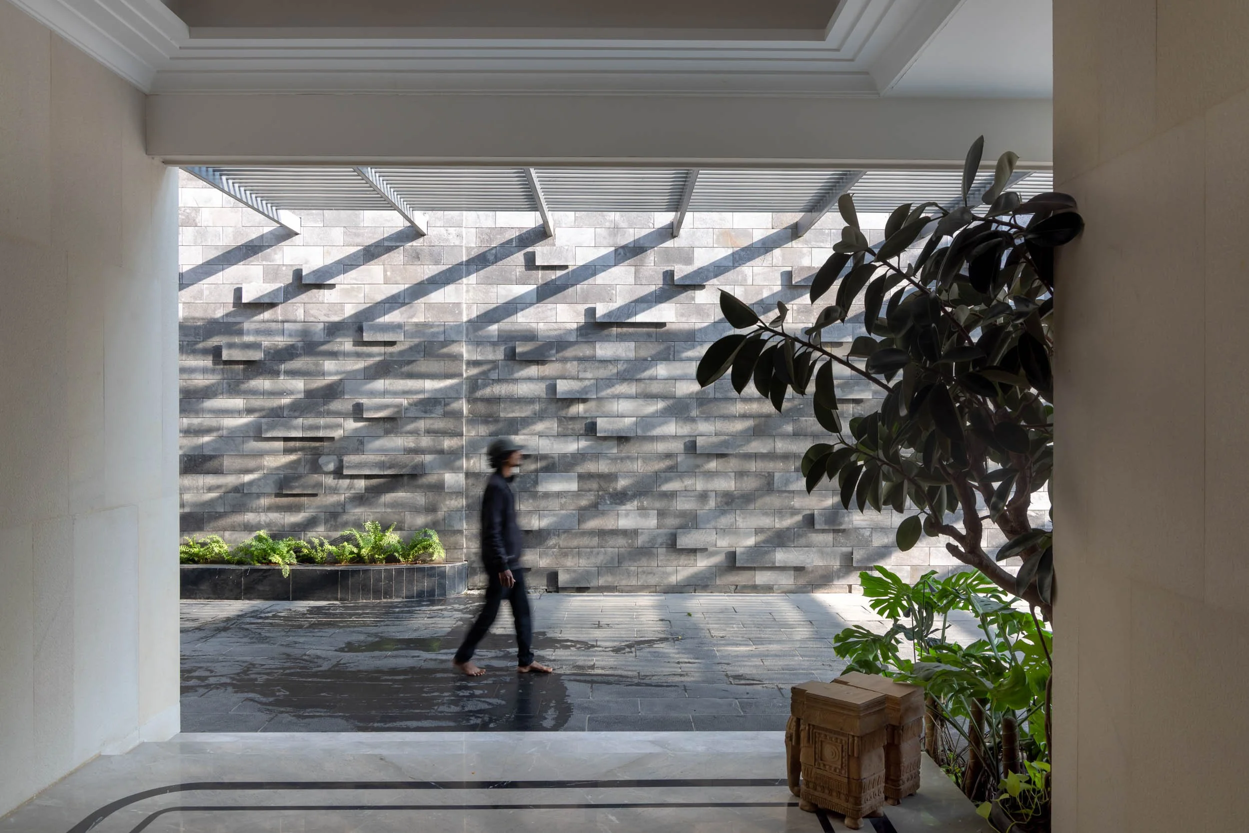 Modern courtyard design featuring a slatted pergola casting geometric shadows on concrete walls, with a silhouetted figure walking past tropical planters.