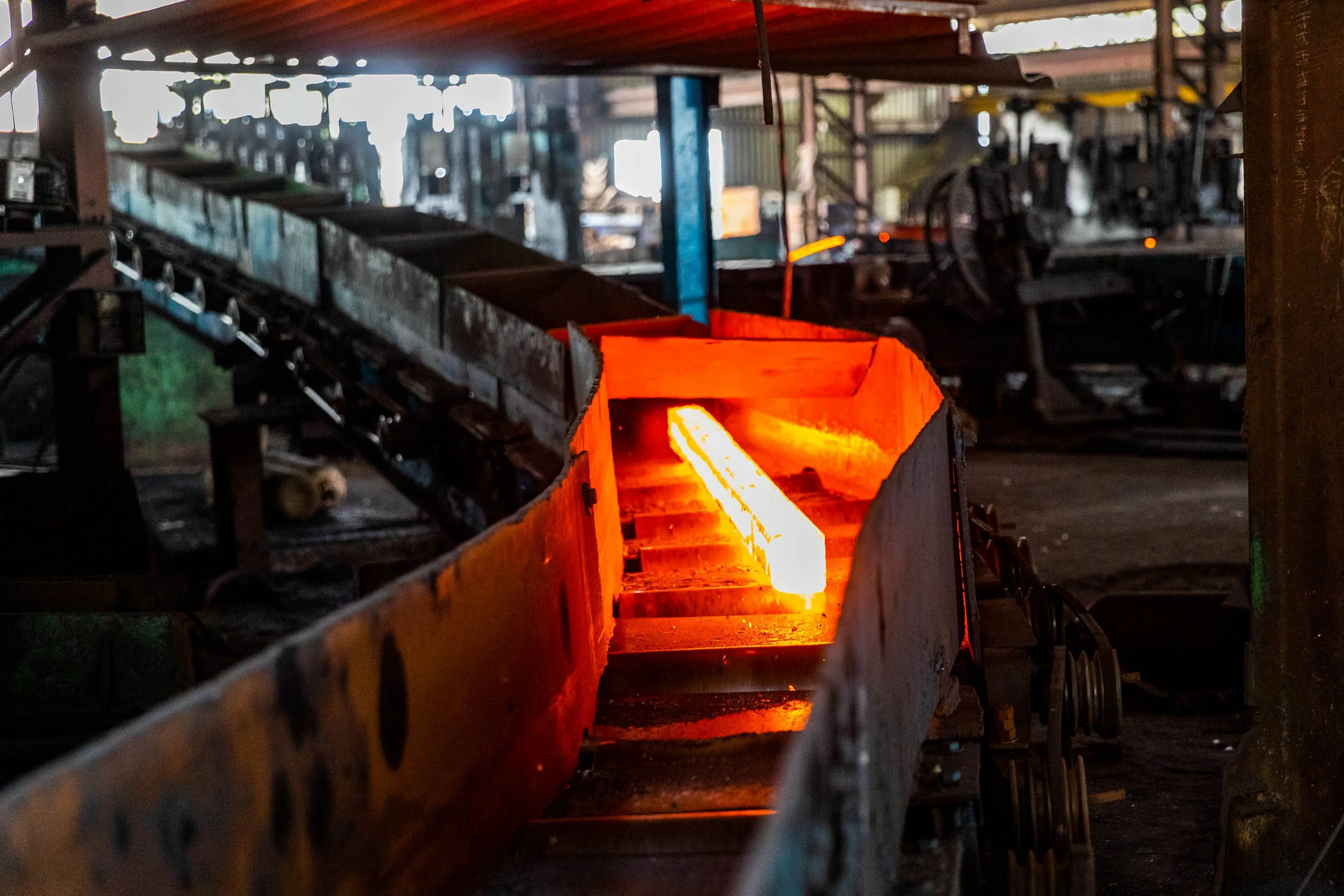 Interior industrial photography of a glowing red-hot steel billet moving along a roller conveyor in a heavy manufacturing plant