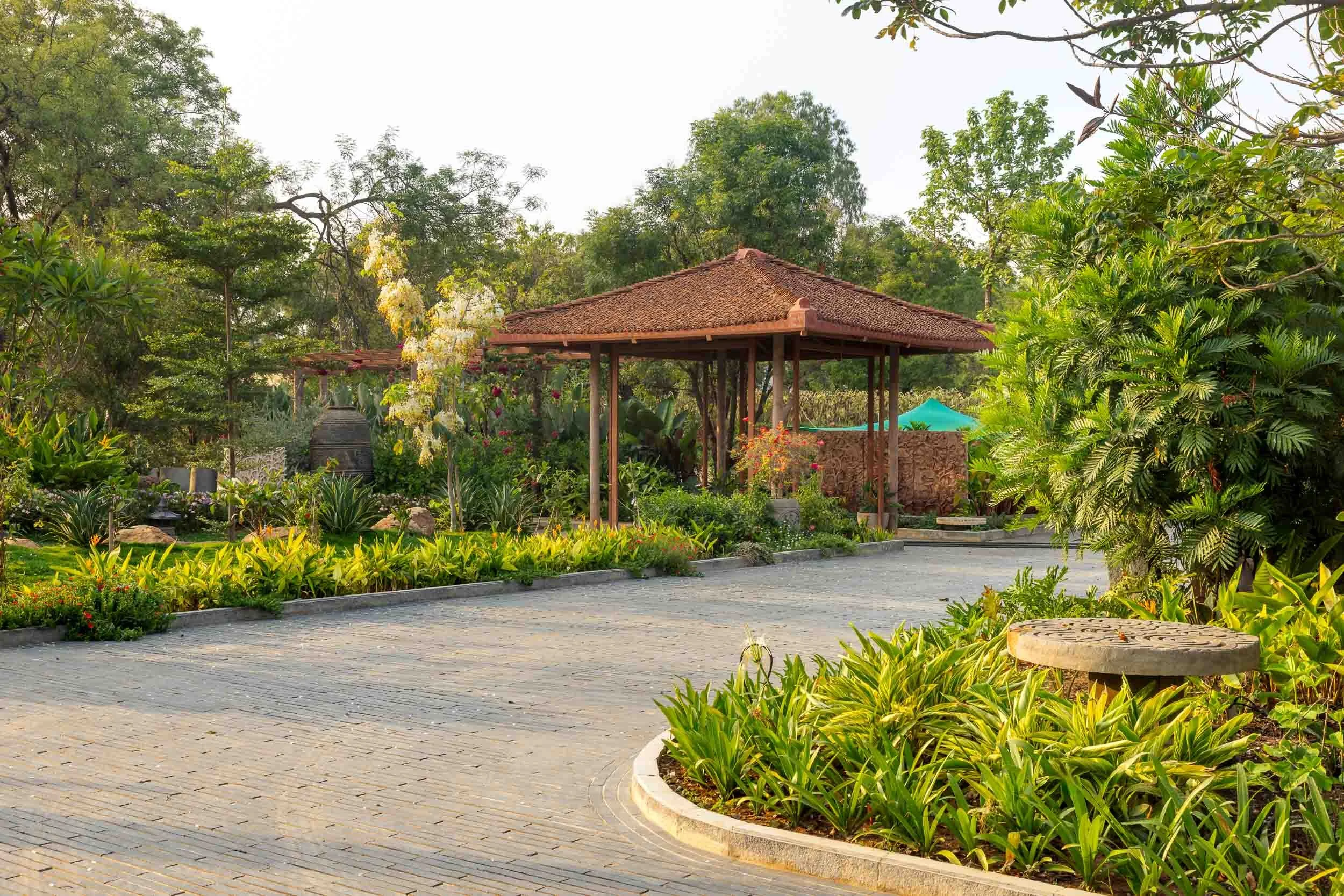 Exterior daytime photography of a luxury resort farm stay featuring a paved circular driveway, vibrant landscaping, and a traditional pitched-roof pavilion