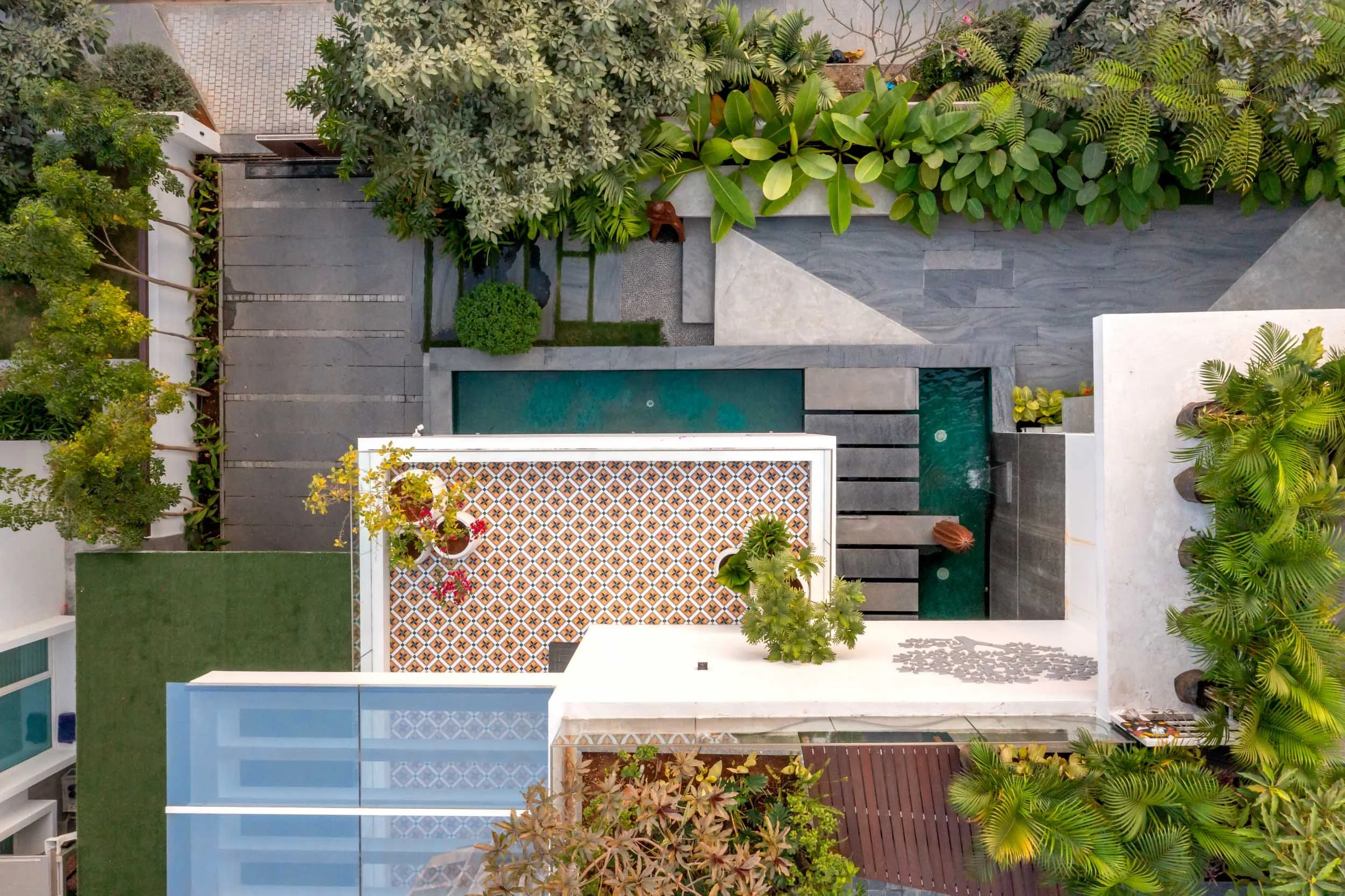 Aerial view of a modern residential backyard featuring a geometric lap pool, white patio spaces, and lush green landscaping