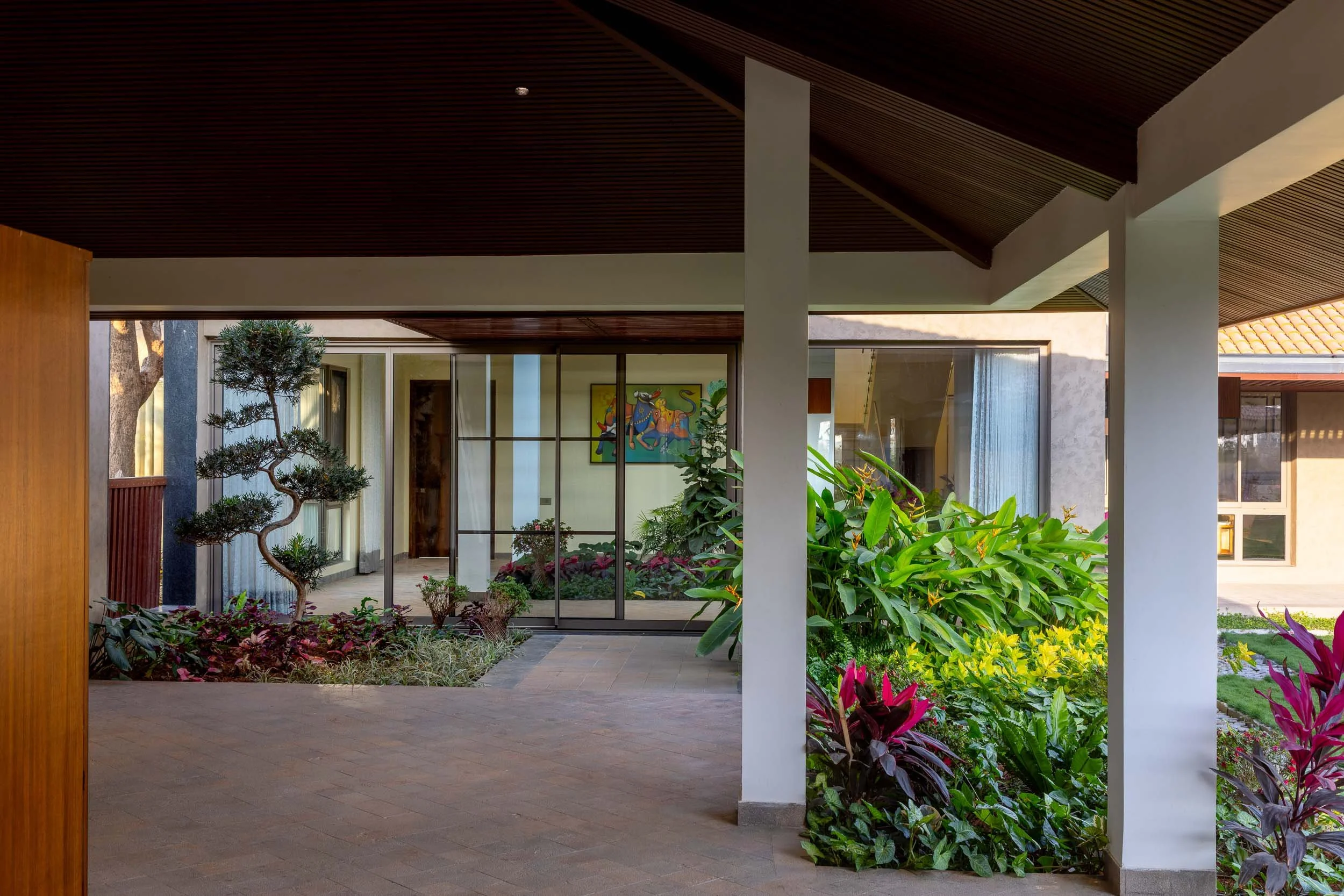 Exterior daytime photography of a covered veranda at a luxury resort featuring a slanted wooden ceiling, white pillars, stone flooring, and lush green potted plants