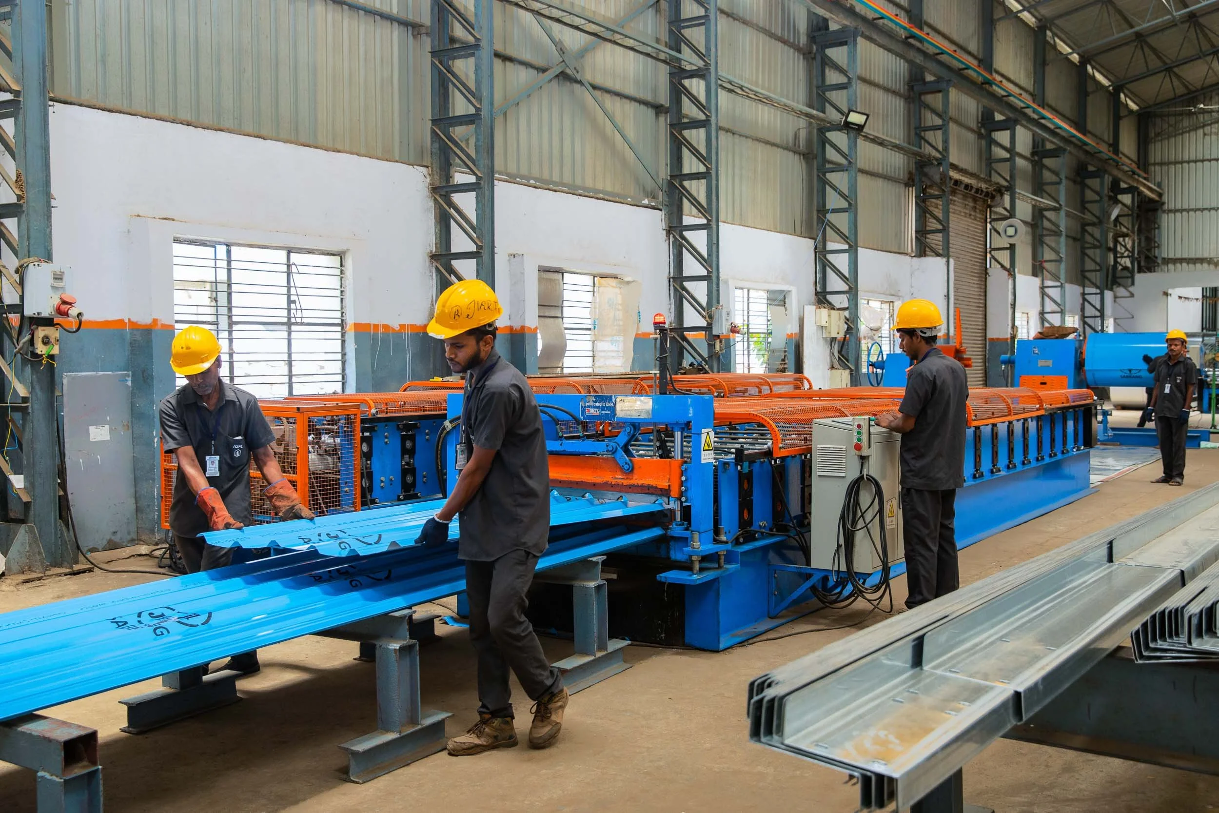 Interior industrial photography of factory workers in yellow hard hats handling a long blue structural material on a production line in a manufacturing plant