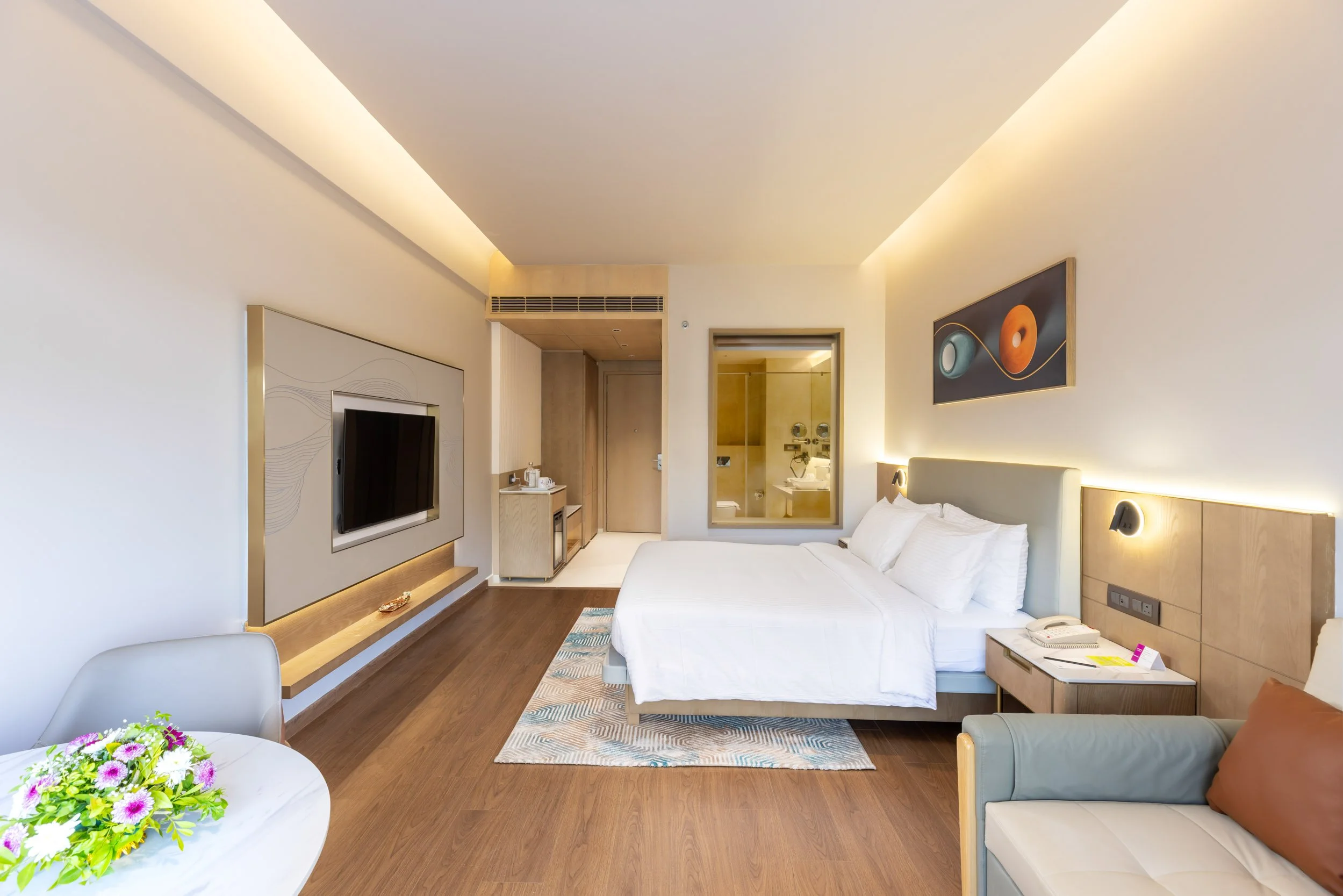 Interior photography of a spacious modern hotel bedroom featuring a king bed, a long integrated wooden desk, a flat-screen television, and a tray ceiling with warm ambient lighting