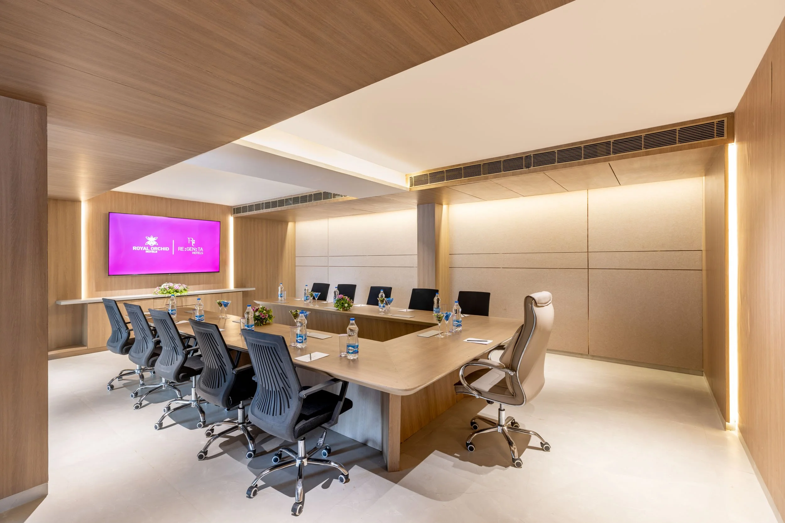 Interior photography of a modern hotel boardroom featuring a long wooden conference table, ergonomic black office chairs, wood-paneled walls, and a large display screen