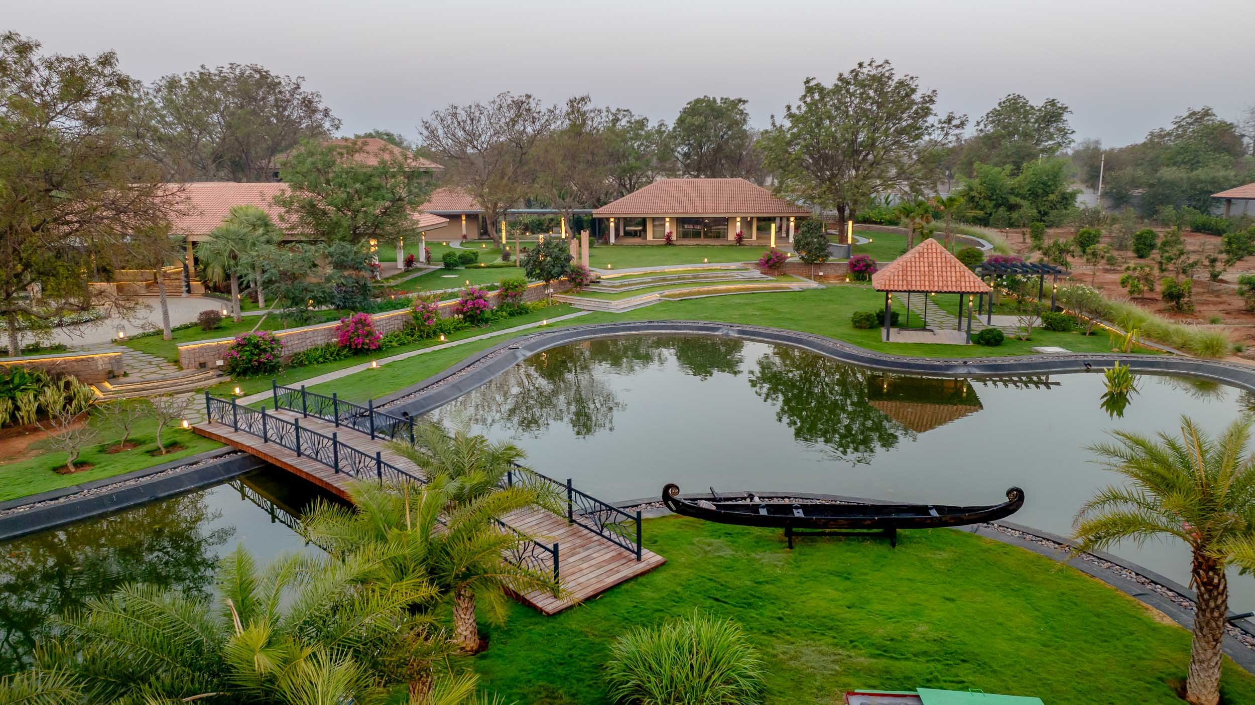 Elevated landscape photography of a luxury resort farm stay featuring a large pond with a traditional wooden boat, manicured lawns, and pitched-roof buildings.