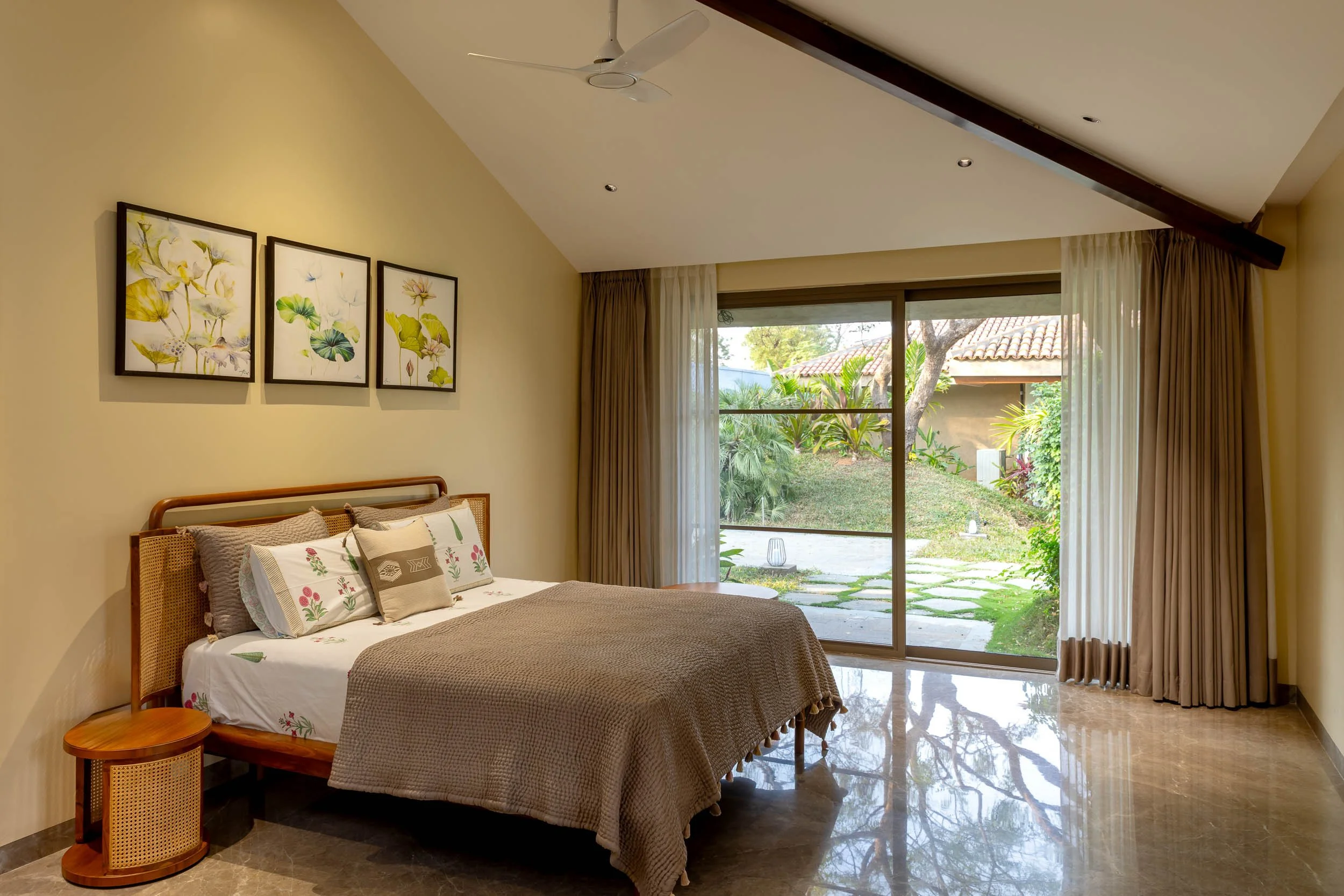 Interior photography of a rustic-modern luxury resort bedroom featuring a pitched wooden ceiling, a large bed, and expansive windows with views of a lush garden.