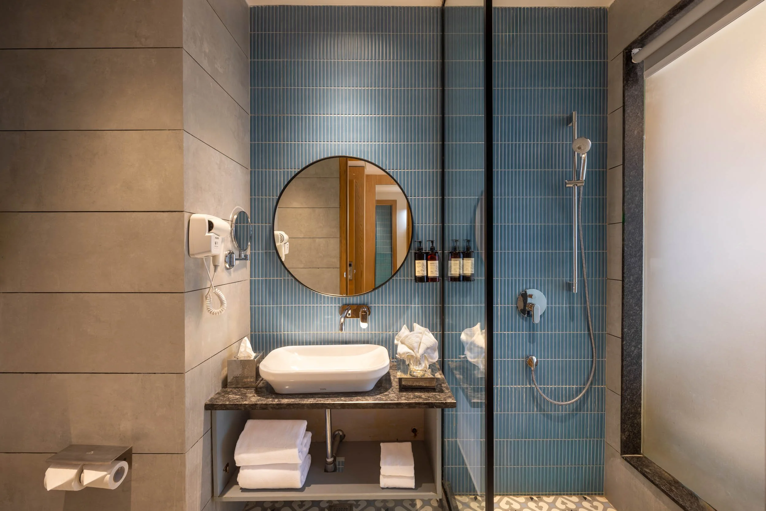 Interior photography of a modern luxury hotel bathroom featuring blue fluted wall tiles, a round backlit mirror, a white washbasin, and a glass-enclosed shower.