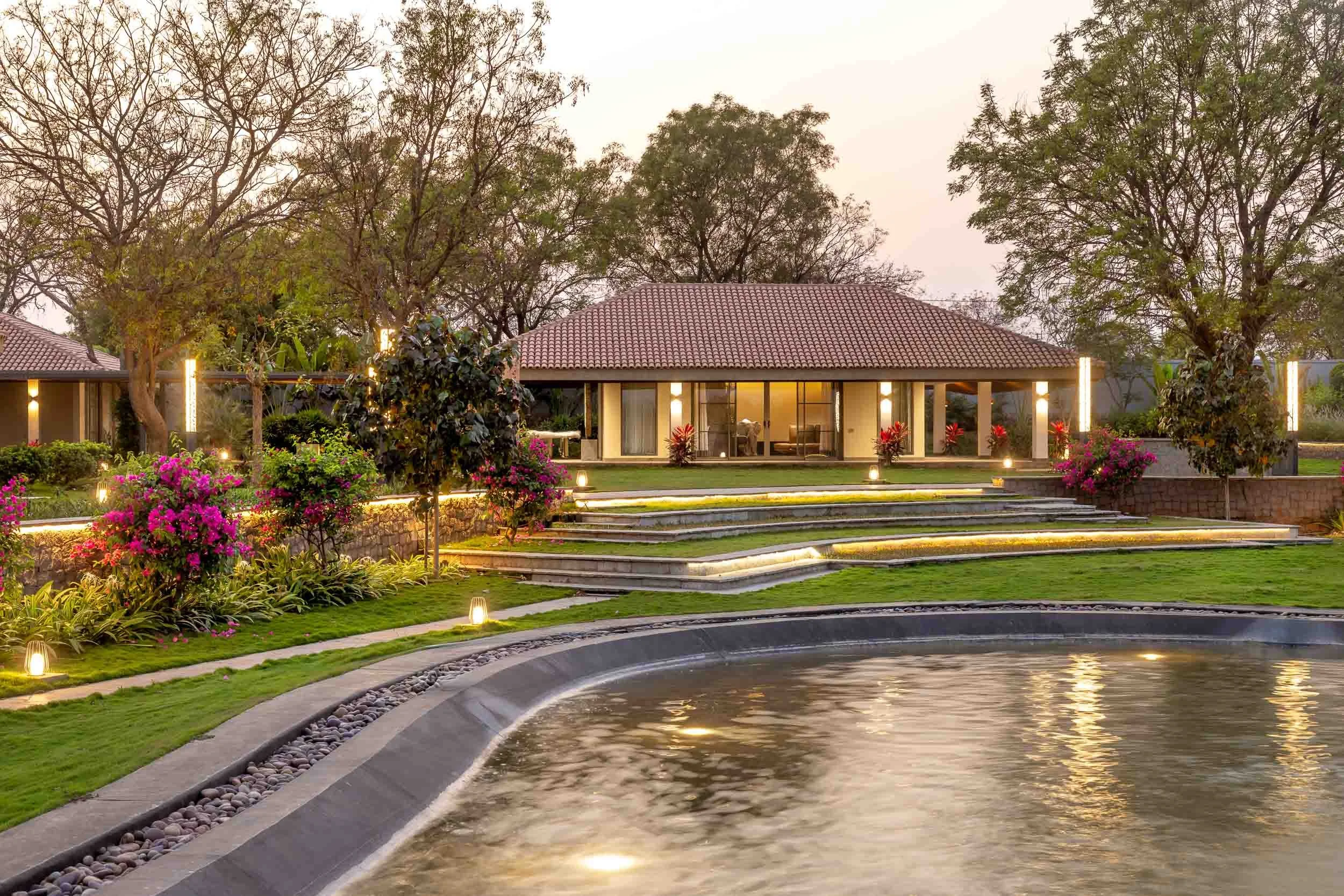 Exterior twilight photography of a luxury resort farm stay featuring warm building lighting, landscaped gardens, and a large reflective pool in the foreground.