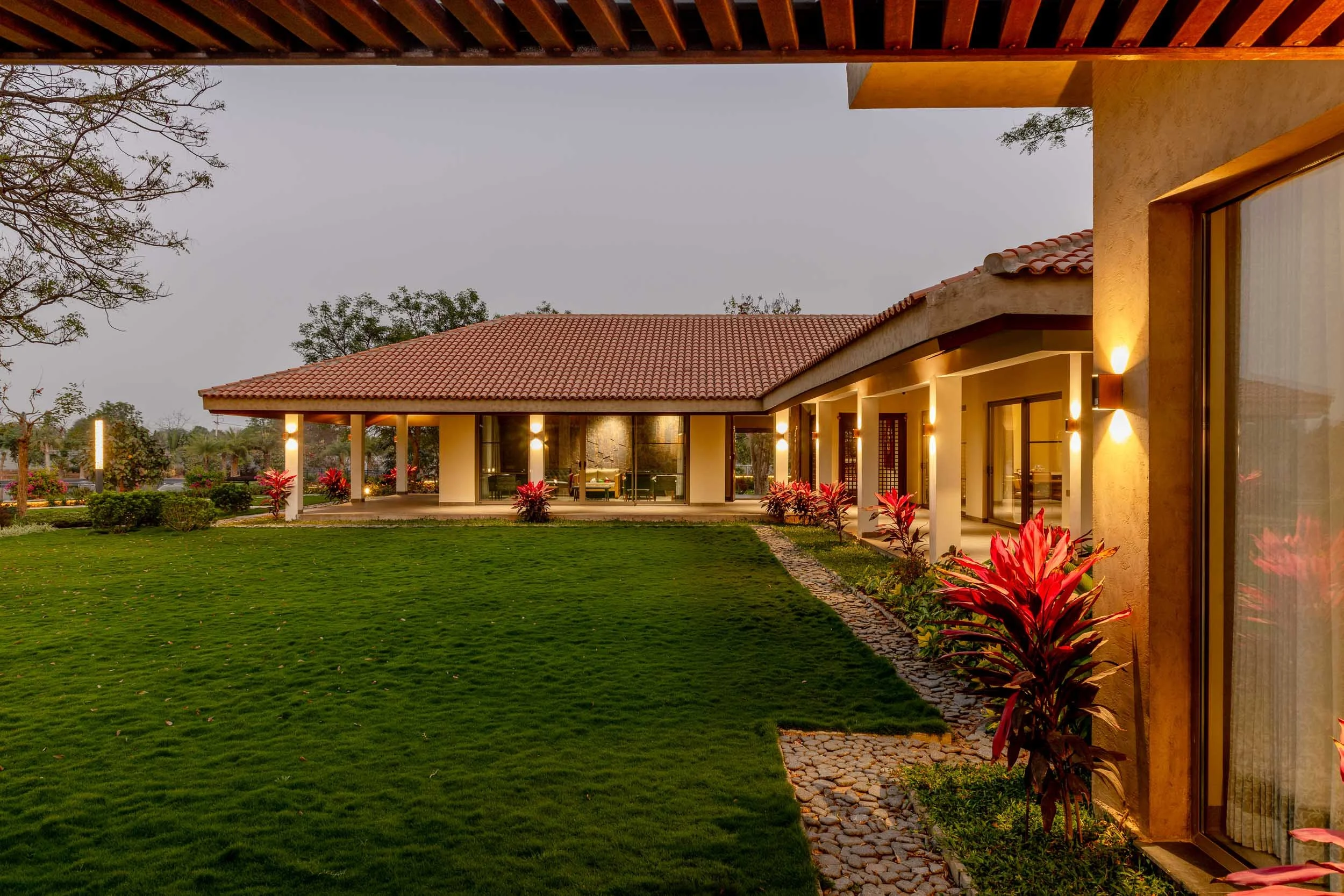 Exterior twilight photography of a luxury resort farm stay featuring a long traditional building with a pitched roof, warm ambient veranda lighting, and an expansive green lawn