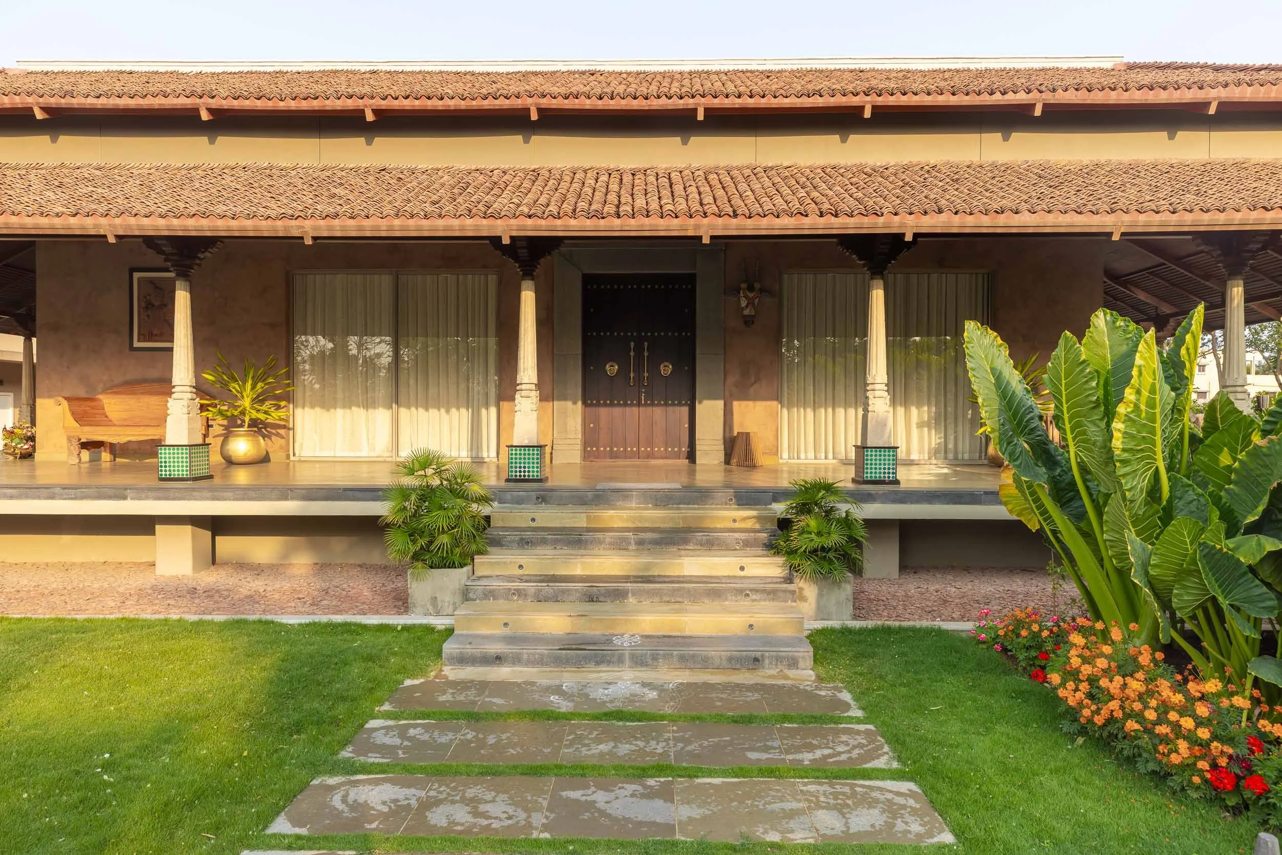 Exterior daytime photography of a traditional resort building featuring a pitched roof, a stone pathway with steps, a covered wooden veranda, and a green lawn